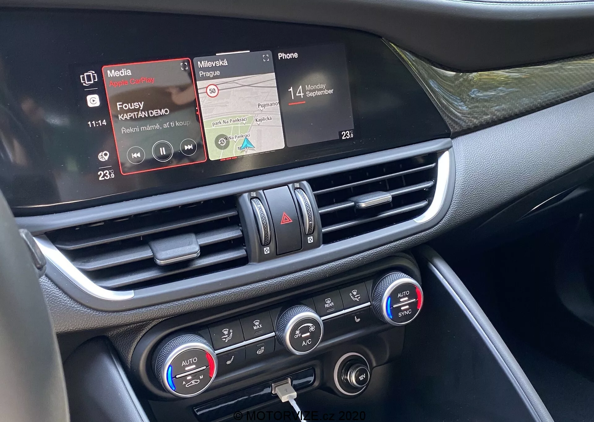 Közelkép a 2016-os Alfa Romeo Giulia középső műszerfali részéről az anyósülés perspektívájából. A közepesen közeli képen kiemelkedik az infotainment érintőképernyős kijelző médialejátszó és telefoncsatlakozó funkciókkal, a krómozott légbeömlő nyílás és a klímavezérlők kétzónás hőmérséklet-beállítással és digitális kijelzőkkel. A műszerfal anyagai között műanyag és fényes vagy krómozott részletek találhatók, bemutatva a jármű fejlett belső technológiáját, amely a csatlakoztatásra, médiakijátszásra és klímakontrollra összpontosít.