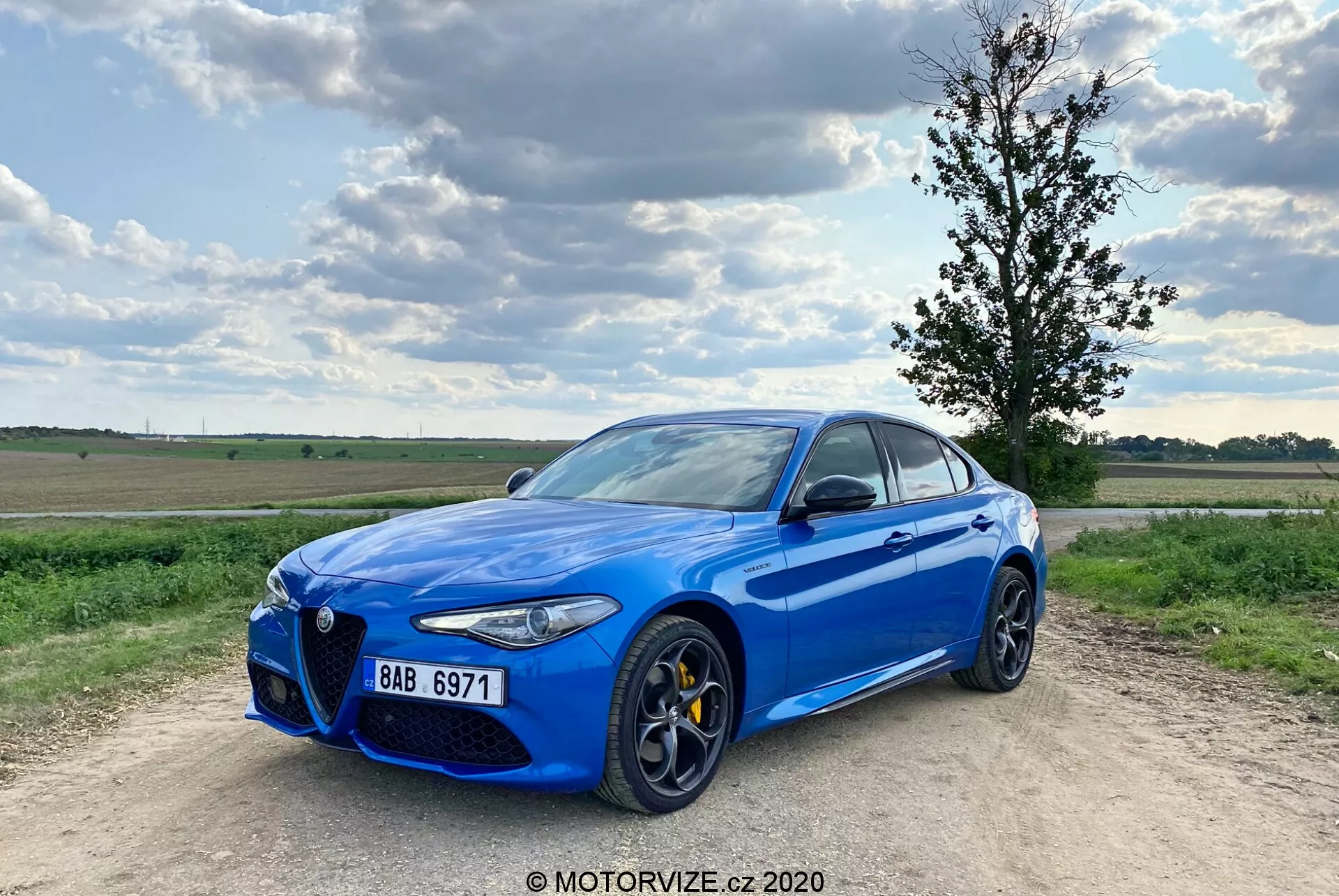 Egy fényes kék színű Alfa Romeo Giulia 2016 sport szedán képe egy kavicsos úton parkolva, részben felhős kék ég háttérrel. Az autót háromnegyed elölnézetben fényképezték, kiemelve annak jellegzetes 'Trilobo' első hűtőrácsát, sárga féknyergeit, nagy többrudas könnyűfém keréktárcsákat, elegáns LED fényszóróit és krómozott ablakkereteit. A jármű kissé balra van eltolva, a bal első kerék kifelé mutat, ami dinamikus állást biztosít számára.