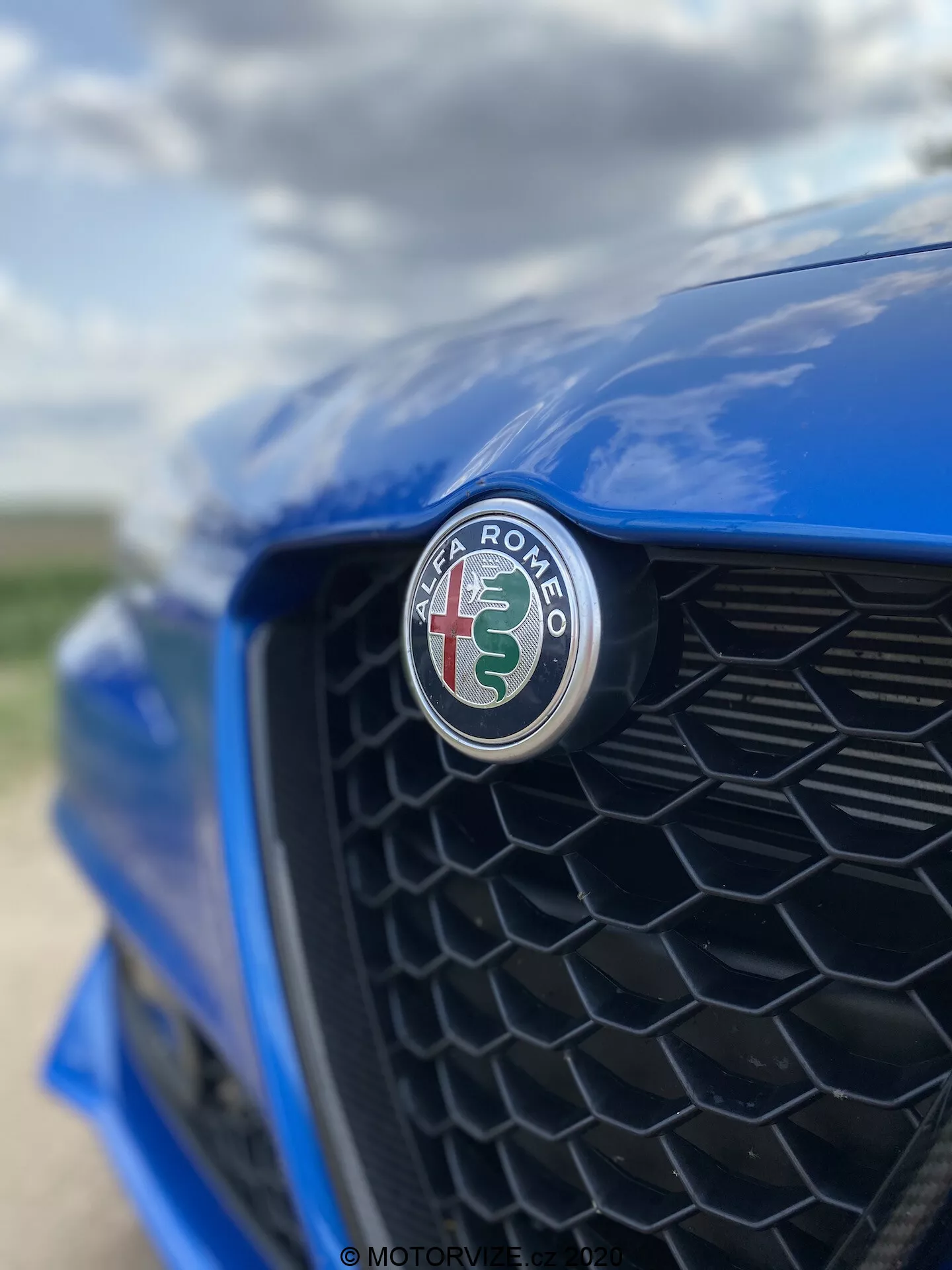 Közelkép egy kék Alfa Romeo Giulia 2016 elülső részéről, a gyártó jelképére összpontosítva, amely vörös keresztet és zöld kígyót ábrázol fehér háttéren. A felvétel egy kissé oldalirányú szögből készült, a vezető oldala felé, a fekete méhsejtmintás hűtőrács enyhén elhomályosodik, kiemelve a jelkép éles fókuszát és a fényes felületet.