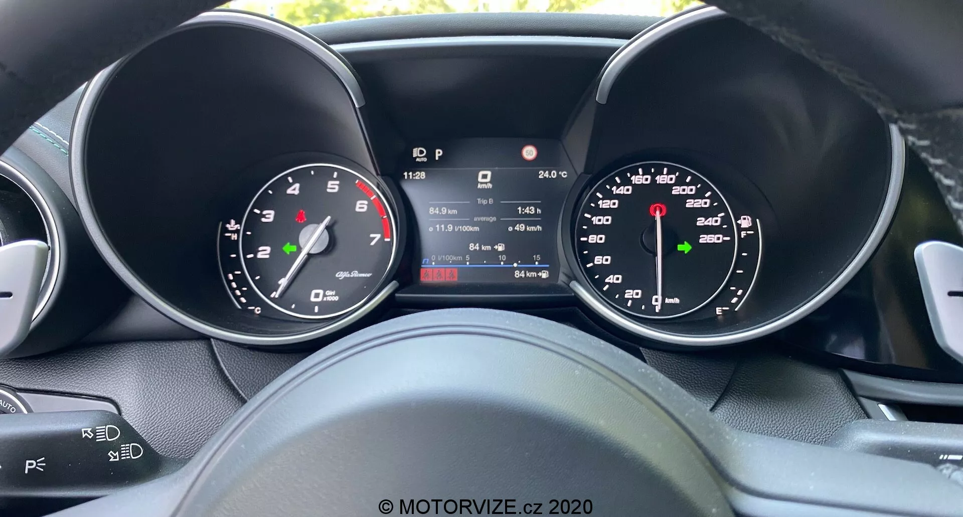 A sofőr nézőpontjából készült kép a 2016-os Alfa Romeo Giulia műszerfaláról, középső digitális kijelzővel, amely mutatja a sebességet, fokozatot, hőmérsékletet és kilométeróra állását, analóg sebességmérő és fordulatszámmérő mérőműszerek mellett, hagyományos dizájn és modern technológia ötvözetét bemutatva, részleges kilátással az audió és tempomat vezérlőkre a kormánykeréken.