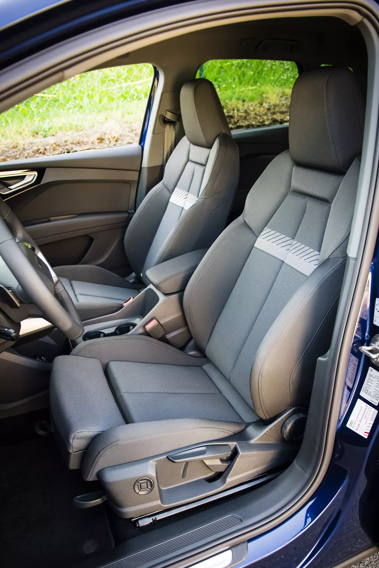 2021-es Audi Q4 e-tron belső terének bemutatása, ahol látható az utasoldali első ülés kétszínű kárpitjával, részletes varrással, elektromos beállítószabályzókkal és biztonsági övvel, mellette a részben látható osztottan hajtható hátsó ülés és a kabin technológiai jellemzői.