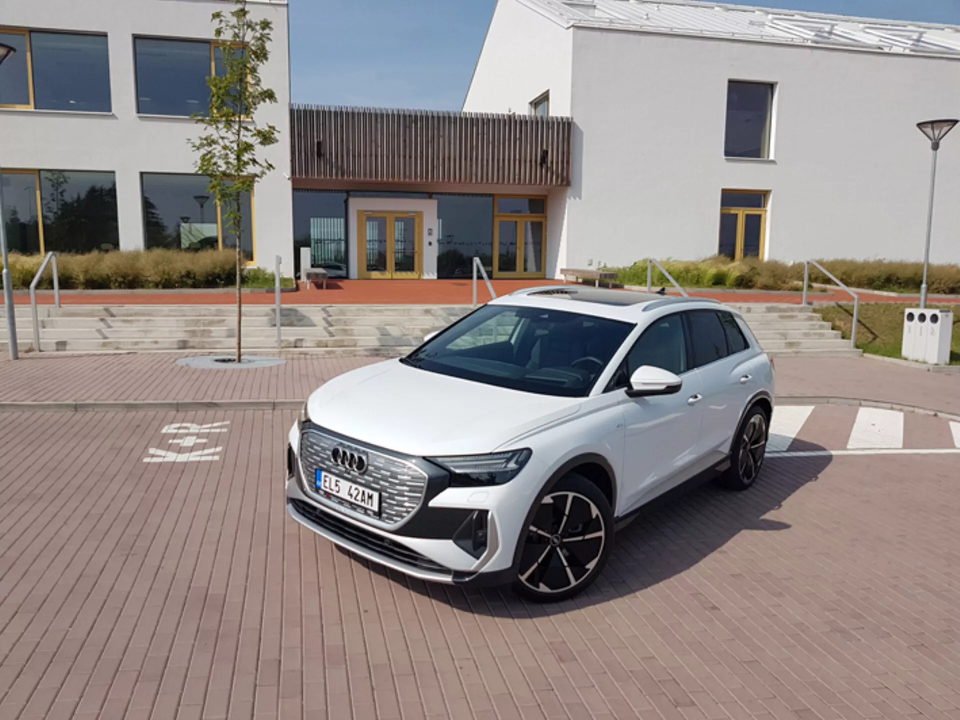 2021-es fehér Audi Q4 e-tron, moderne LED-es fényszórókkal, hatszögletű első hűtőráccsal és nagy ötvözött kerekekkel, téglamintás burkolaton parkol, városi építészettel és egy töltőállomással a háttérben, tiszta déli égbolt alatt.