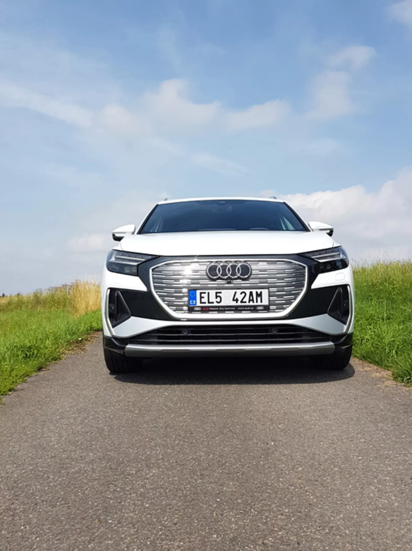 2021-es Audi Q4 e-tron középre igazított első nézete, amely nagy méretű hatszöges hűtőrácsot ábrázol Audi emblémával, LED fényszórókat, európai rendszámtáblát, lehetséges ADAS szenzorokat, légbeömlőket, fémes fényezést és króm díszítéseket részben felhős ég alatt, aszfaltozott úton füves oldalakkal.