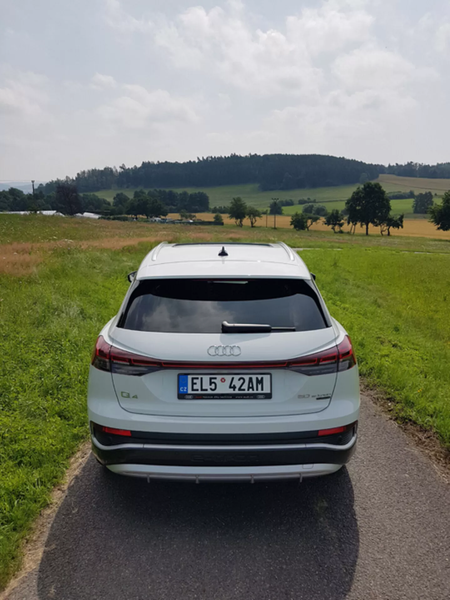Egy ezüst Audi Q4 e-tron 2021 hátsó nézete egy keskeny, burkolt úton, pásztori tájkép közepette mezőkkel és fákkal részben felhős ég alatt, kiemelve elegáns designját, fékfény sávját, tetősárvédőjét, hátsó parkoló szenzorait és kéttónusú ötvözött kerekeit.