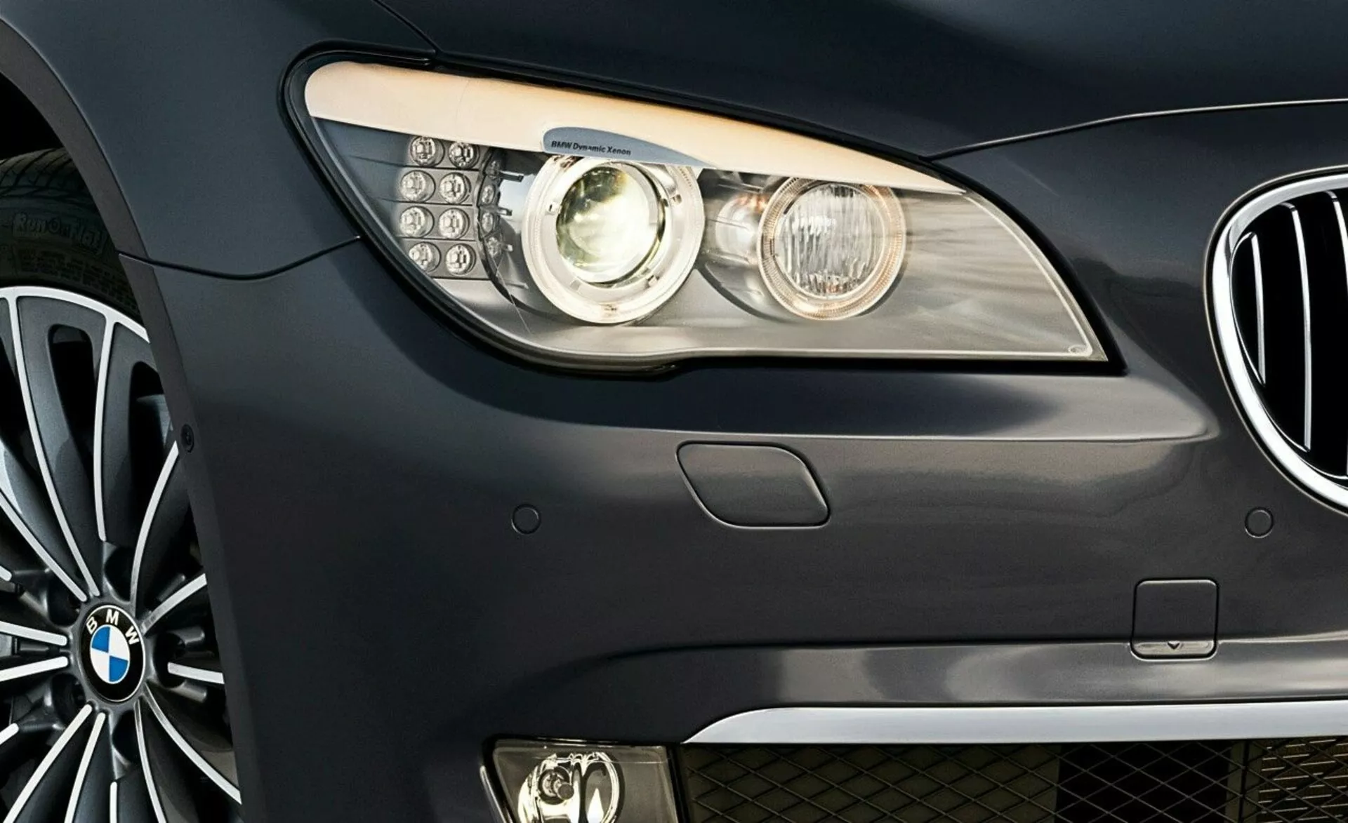 Közeli oldalnézet a 2005-ös BMW 7-es sorozatú első részéről, a márka emblémájával, modern fényszóró-egységgel projektor lencsével és LED nappali menetfényekkel, részleges nézet a vesemedence-rácsról és a többsugaras ötvözött kerékről.