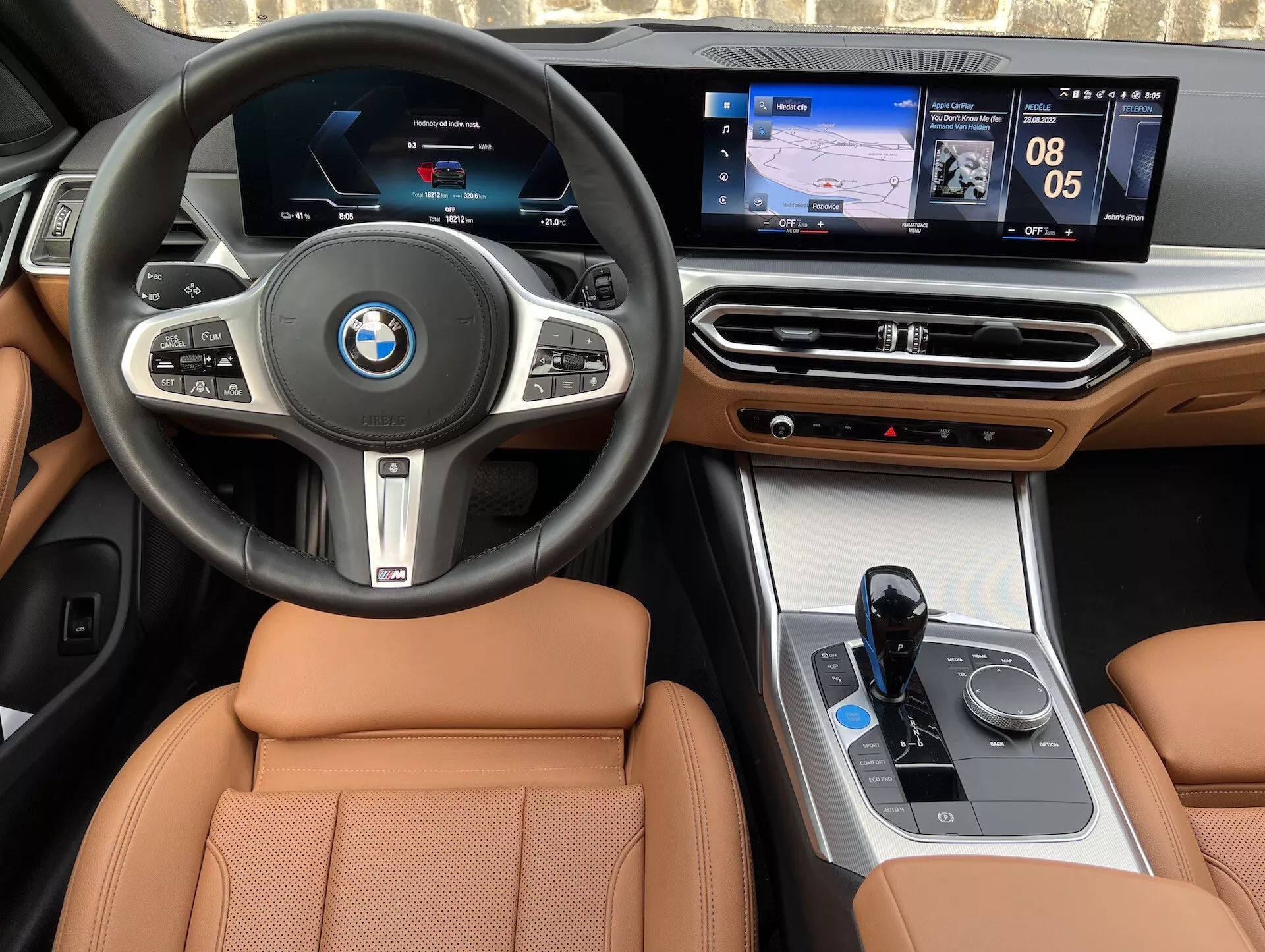2021-es BMW i4 belső nézete, központi kormánykerékkel BMW logóval, multifunkciós gombokkal, digitális műszerfallal, érintőképernyős központi infotainment rendszerrel, amely navigációt és médiát jelenít meg, luxus fa díszítésekkel a műszerfalon, klímavezérlő interfésszel, elektronikus váltóval, iDrive vezérlővel és barna bőr ülésekkel.