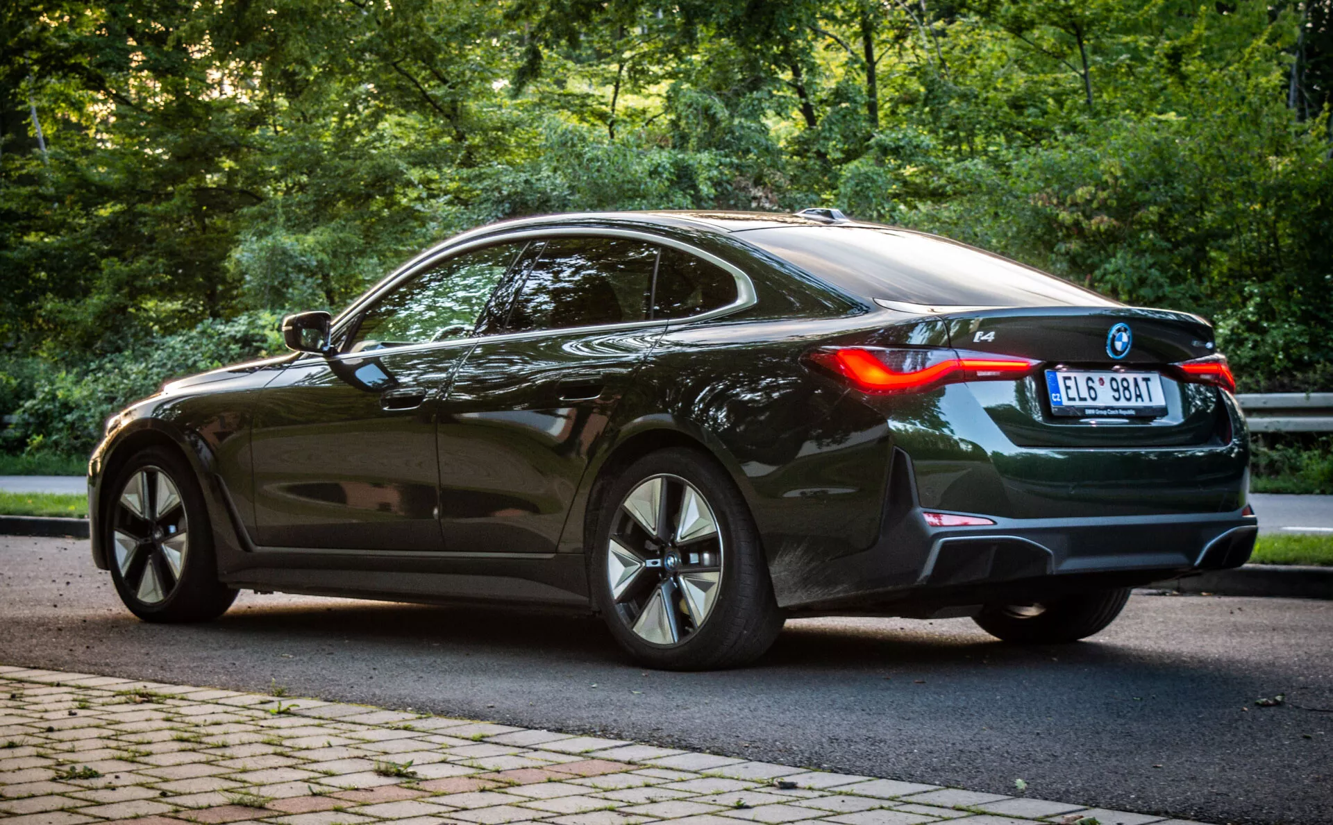 Fényes fekete 2021-es BMW i4 kupé háromnegyedes hátsó nézetben, sportos többsugárzós ötvözött kerekekkel, LED hátsó lámpákkal, és panoráma napfénytetővel, zöld, fás környezetben, nappali fényben