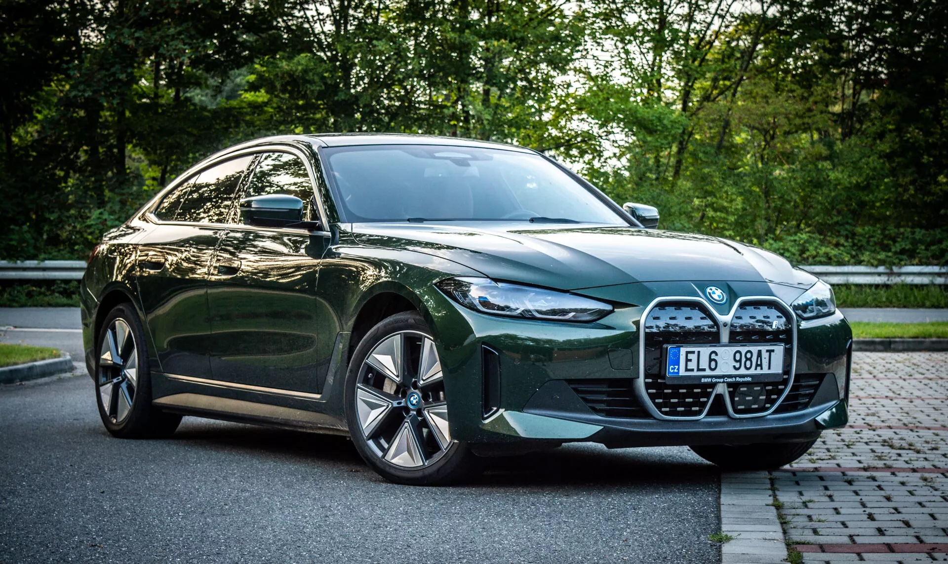 2021-es zöld BMW i4 négyajtós szedán parkol téglaburkolatú utcán, füves terület mellett, háttérben lombozattal, az autó első negyedét bemutató nézőponttal, amely kiemeli a nagy hűtőrácsot, LED fényszórókat, légbeömlőket és ötvözött kerekeket