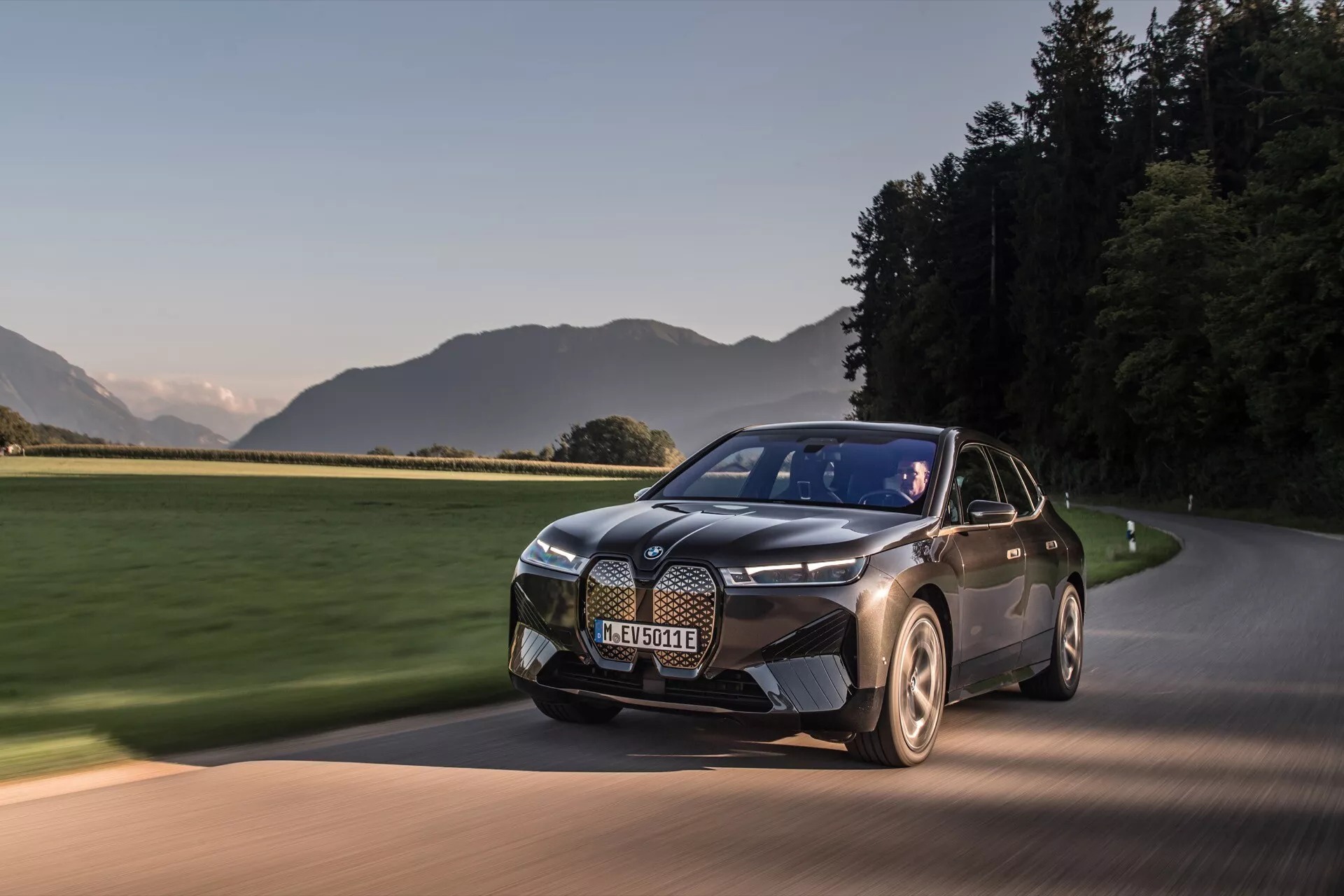 Modern BMW iX 2021 (i20) elektromos SUV mozgásban egy aszfaltozott úton hajnalban vagy alkonyatkor, bemutatva a kéttonusú festését, elegáns tervezését kiemelkedő rácsával, különleges fényszóróival és aerodinamikus kerekeivel egy festői háttér előtt.