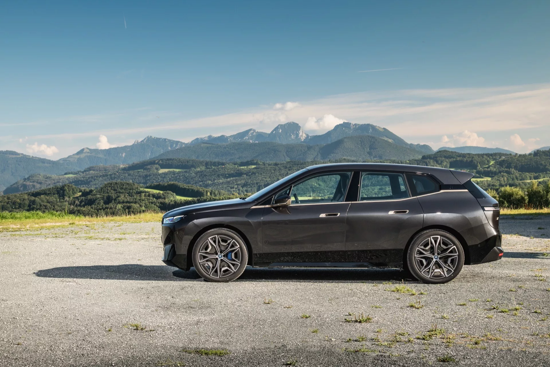 2021-es BMW iX (i20) modern SUV oldalnézete kavicsos talajon hegyvidéki háttérrel tiszta kék ég alatt, nagy méretű többsugaras öntvénykerékekkel, elegáns tervezésű magas övvonallal és lejtő hátsó tetővonallal, személyiségi jogot védő üvegezéssel, karosszéria színű tükrökbe integrált irányjelzőkkel, látható első hűtőráccsal és LED fényszóró csoporttal, krómozott díszlécekkel és panoráma tetővel.