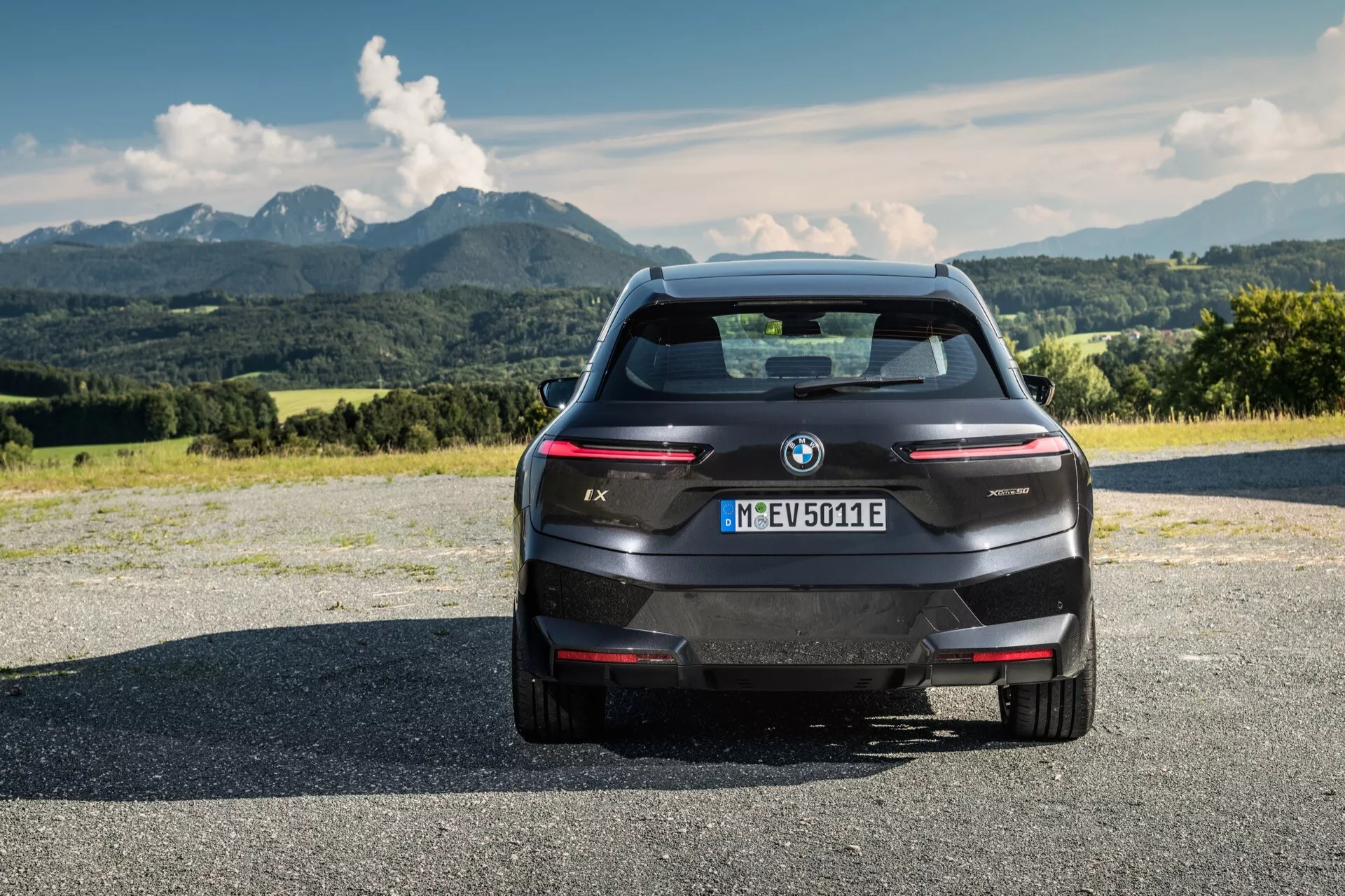 BMW iX 2021 elektromos SUV hátsó nézete aszfaltfelületen, hátterében hegyekkel és részben felhős égbolttal, bemutatva a modern tervezését, elegáns vonalait, a BMW logót és a modell megjelölését, integrált hátsó parkolóérzékelőket, magasra szerelt stoplámpát, hátsó kamerát, vékony, vízszintes hátsó lámpákat és aerodinamikus hátsó ablakot a hagyományos kipufogó hiányával, jelezve az elektromos természetét és a müncheni regisztrációt.