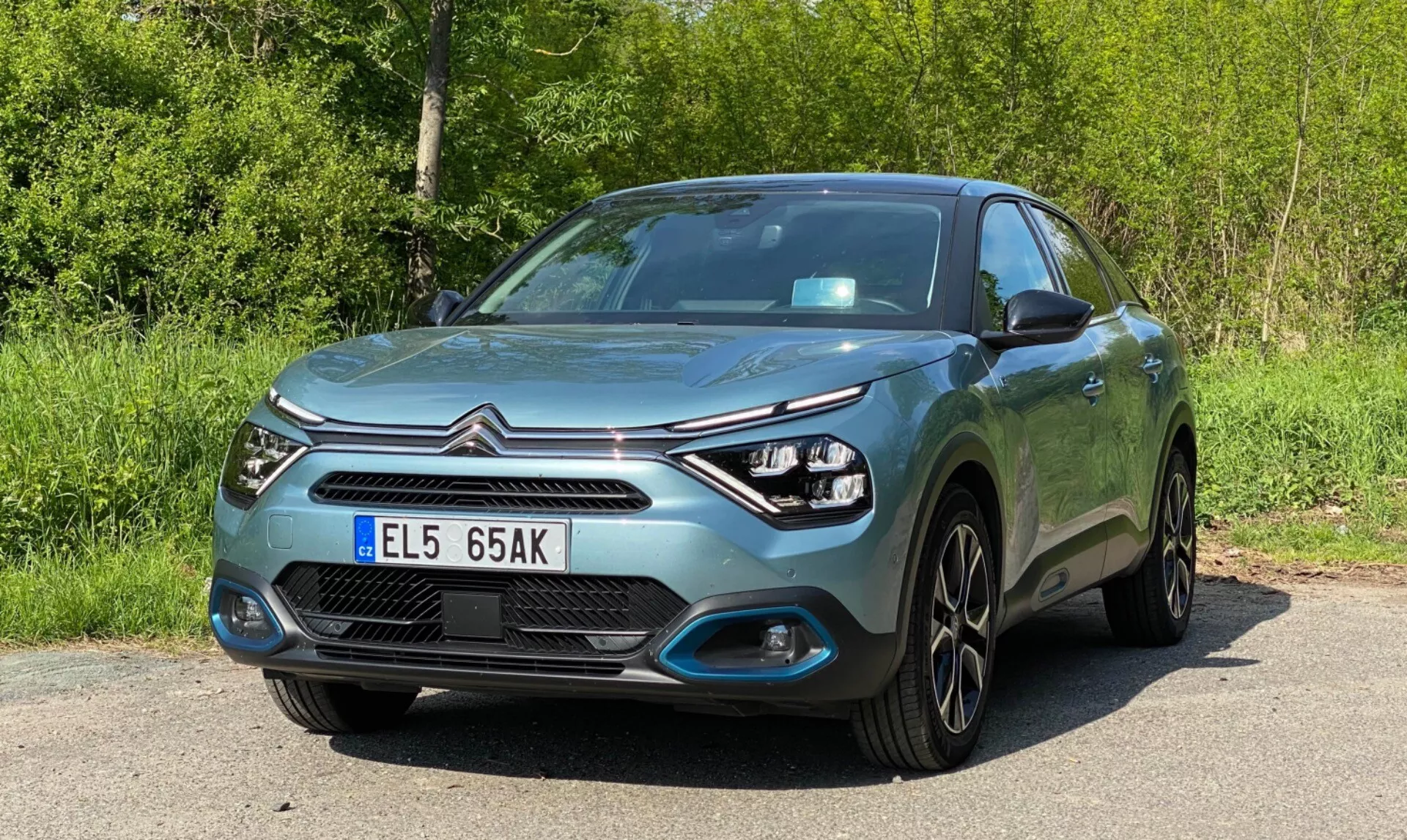 A 2020-as Citroen C4 (E-C4) modern ferdehátú autó alacsony szögből, elölről háromnegyedes nézetben látható a képen, bemutatva az autó elejét, a vezető oldalát, a motorháztetőt, a fényszórókat és a kerék kialakítását. A jellemzők közé tartozik a LED-es nappali menetfény az elegáns fényszóró egységek körül, a lökhárítóba integrált ködlámpák, a gyártói logóval ellátott központi hűtőrács, krómdíszítésekkel szegélyezve, sima motorháztető vonalak, az autó karosszéria színéhez illő ajtókilincsek és visszapillantó tükrök, többrudas könnyűfém keréktárcsák, metálfény és elülső rendszámtábla. Az autó az út szélén parkol, zöld lomb van a háttérben.