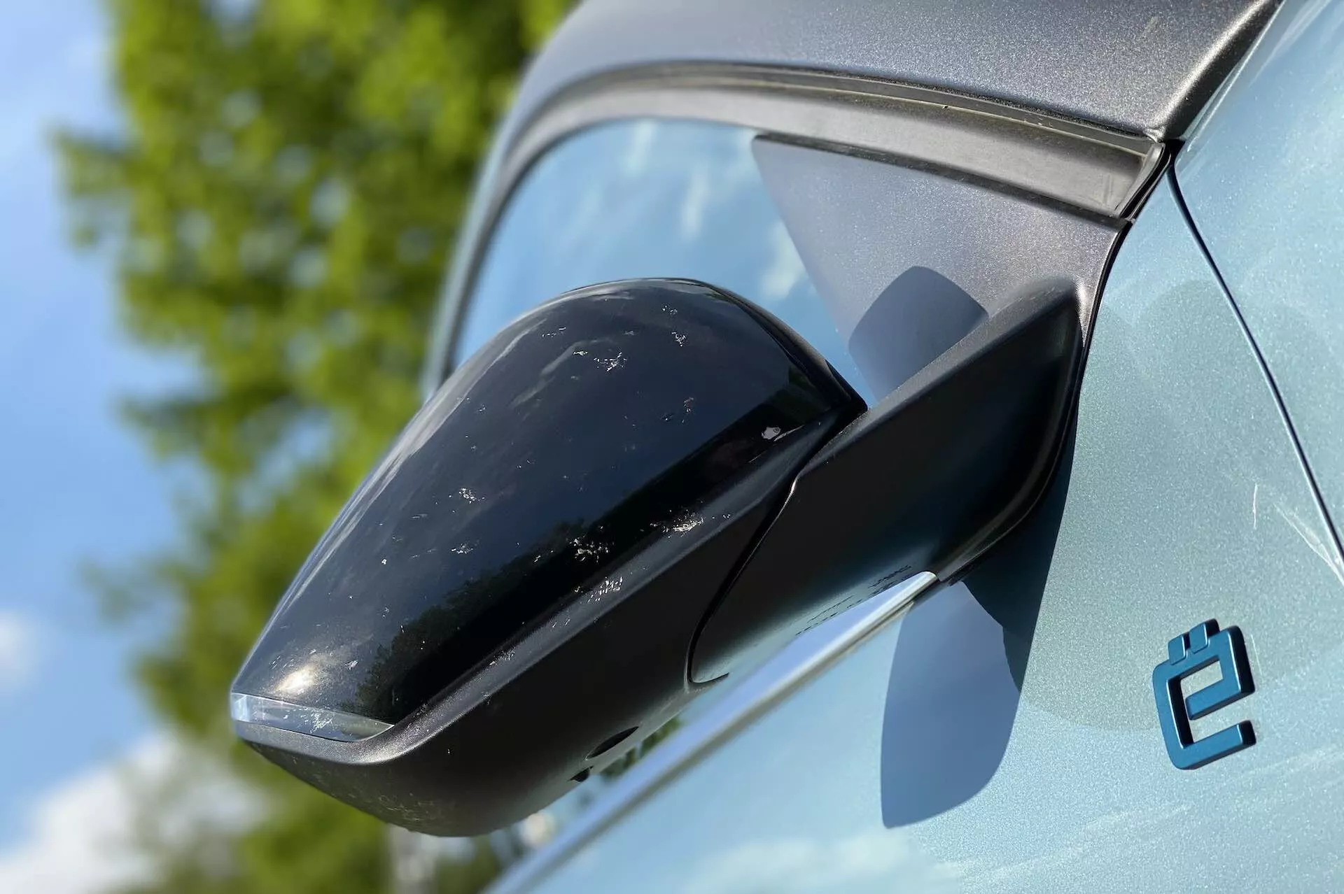 Közelkép a 2020-as Citroen C4 (E-C4) oldalsó tükréről, kiemelve a fekete tükörházat és a világos színű karosszéria kontrasztját. A háttér tiszta égbolt és növényzet látható, utalva a kültéri környezetre. Az autó állapota új, bár a tükörházon némi szennyeződés található. Egy stilizált 'e' embléma is látható, ami valószínűleg egy elektromos változatra utal.