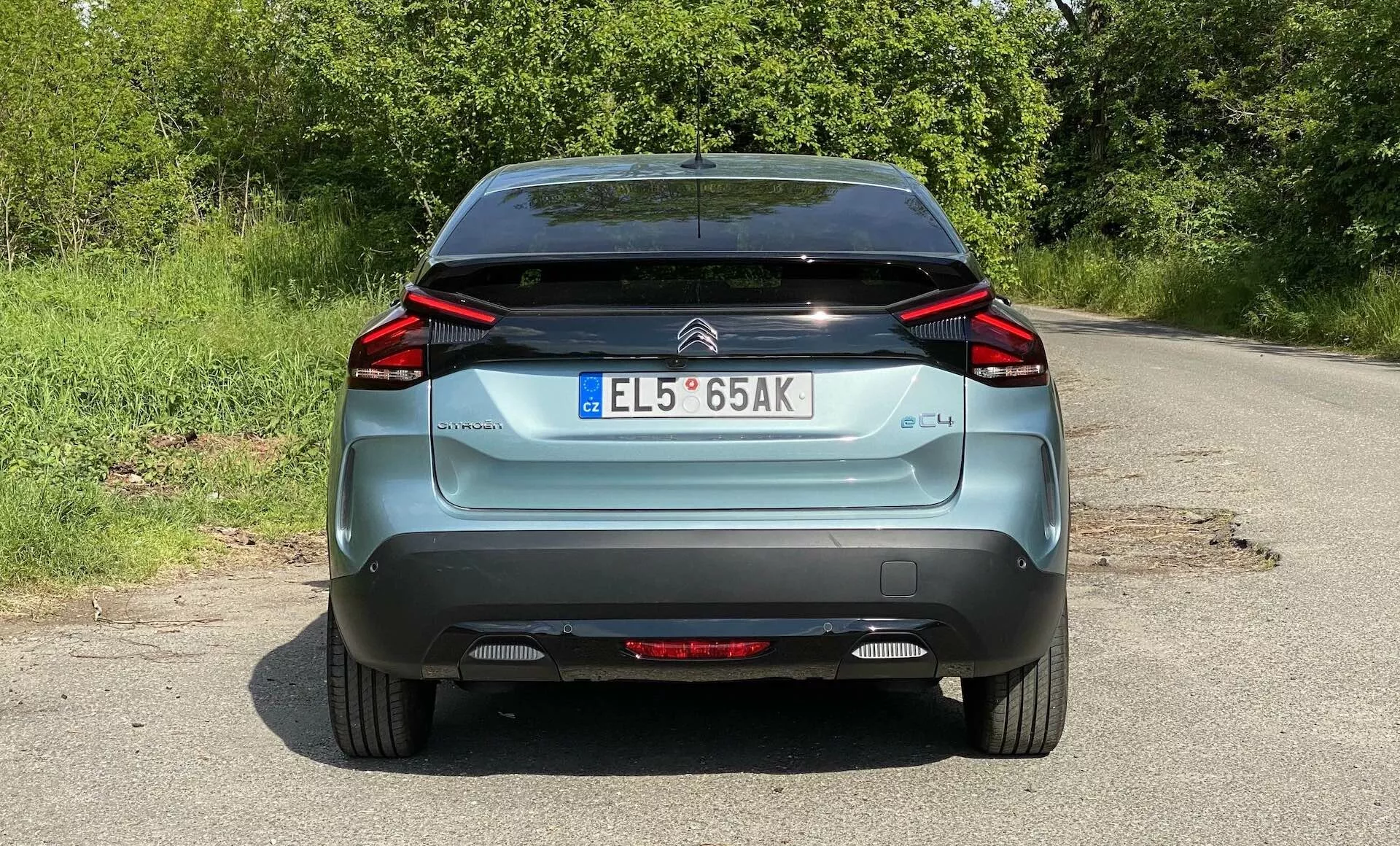Egy kék 2020-as Citroen C4 (e-C4) hátsó nézete, egy vidéki úton parkolva. A kép a jármű teljes szélességét lefedi a kerekek aljától a tetőig, megmutatva a jellegzetes szögletes LED hátsó lámpákat, a márkajelzést, a 'C4' címkét, az európai stílusú rendszámtáblát, a hátsó szélvédőtörlőt és a fekete alsó lökhárító részt. Láthatóak még a reflektorok és a hátsó ködlámpa is.