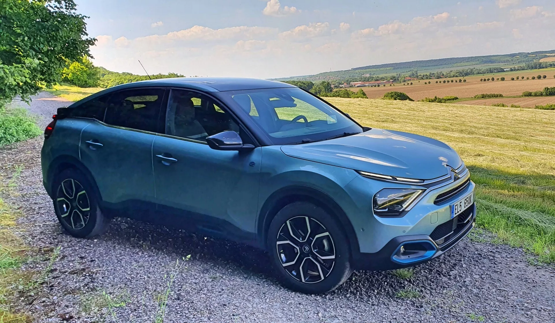 A képen egy Citroen C4 2020 (e-C4) látható oldalról és elölről fotózva, kavicsos úton vagy út mentén parkolva, tájképi háttérrel, enyhe lejtőkkel és tágas éggel. A modern crossover SUV megnövelt hasmagassággal, kerékjárati ívtakarókkal, jellegzetes LED-fényszórókkal, minimalista hűtőráccsal, stílusos többküllős kerekekkel és tompa kék karosszériával fekete elemekkel rendelkezik. A tervezés elegáns és aerodinamikus, sportos testhelyzettel, ami olyan kategóriákat jelez, mint az SUV, Modern Autók, Crossover Járművek, Járműtervezés, Autóipari Technológia és potenciálisan Elektromos Járművek.