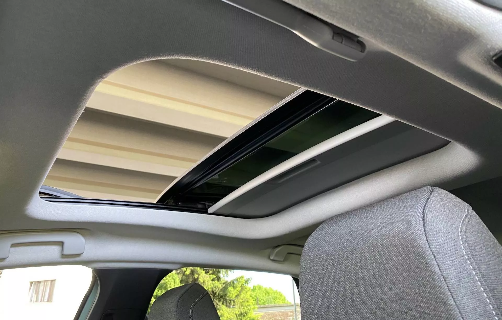 Belső nézet a hátsó ülésről egy 2020-as Citroen C4 (e-C4) esetében, alacsony, kissé balra eltolódott szögből fókuszálva a nyitott napfénytetőre. Az üvegpaneles napfénytető meg van döntve és nyitva van, lehetővé téve a természetes fény beáramlását, amely megvilágítja a világosszürke vagy bézs szövet borítást és az illeszkedő utas ülésének fejtámláját. A kép megörökíti a légiességet és az autó belső tervezésének lényeges részleteit.