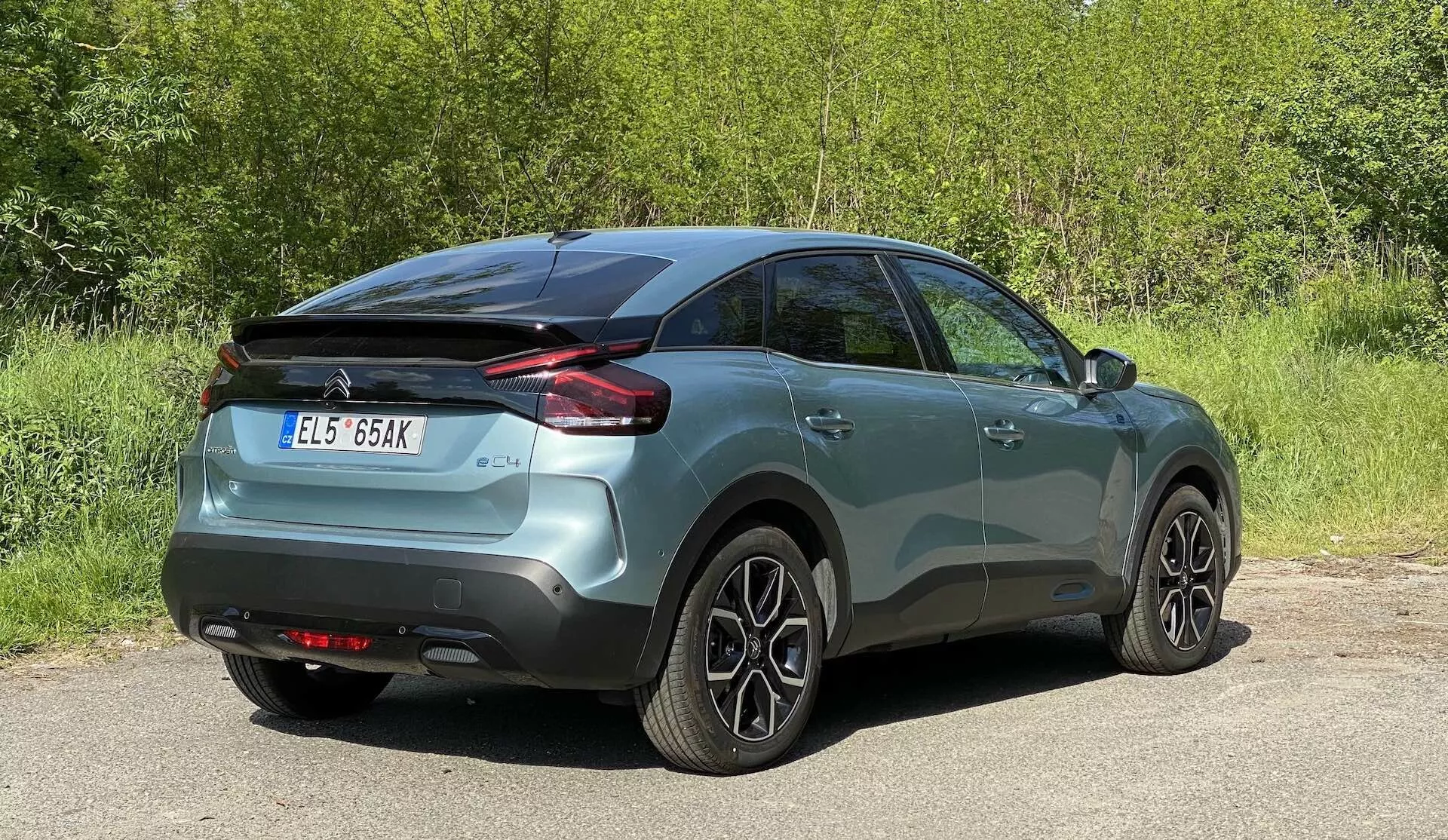 A képen egy Citroen C4 2020 (e-C4) autó látható, amely a hátulsó háromnegyed nézetet mutatja. A fémesen festett jármű központi helyzetben van, oldal- és hátprofilját feltárva, beleértve a vízszintesen tervezett hátsó lámpákat, a karosszéria színű ajtókilincseket és a többküllős könnyűfém kerekeket. A környezet kültéri helyszínt sugall, némi növényzettel a háttérben.