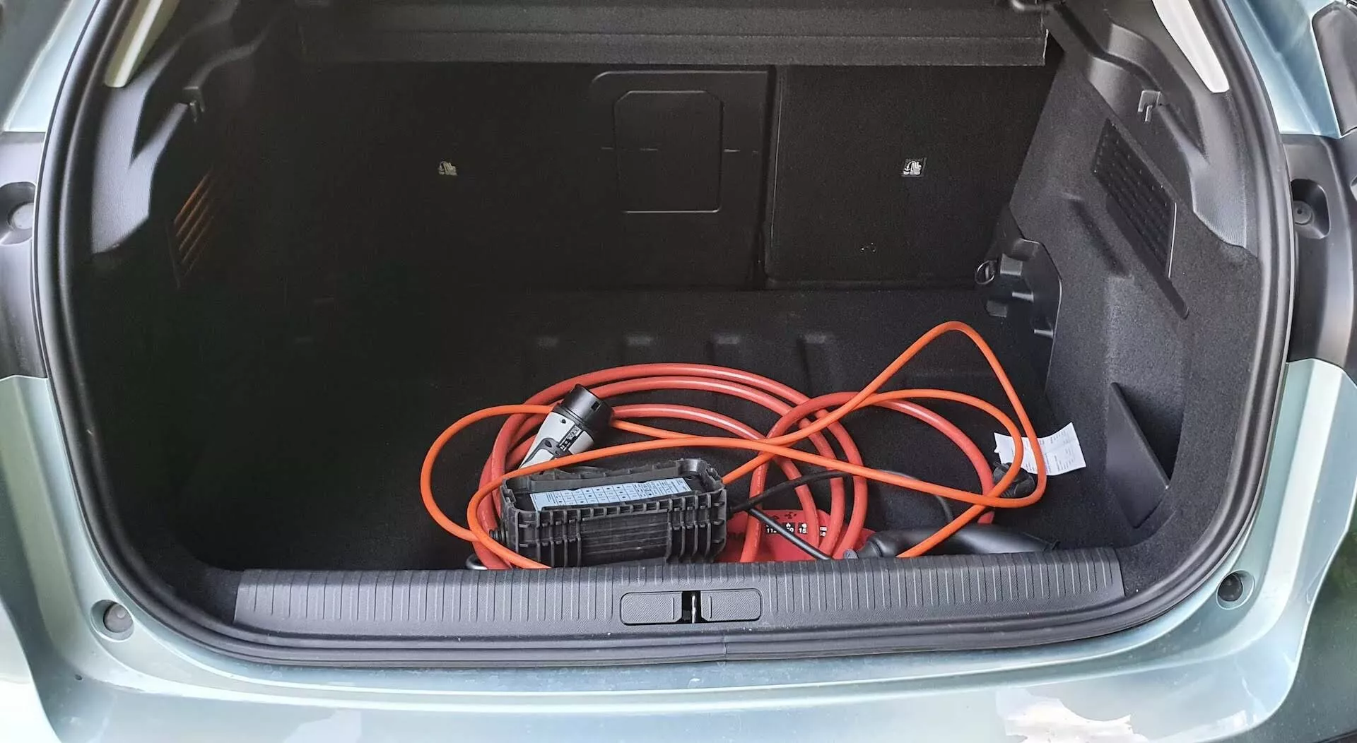 Felülnézet a Citroen C4 2020 (e-C4) ferdehátú csomagtartójára, szőnyeggel borított padlóval, oldalsó panelekkel hozzáférési panellel, és lazán elhelyezett narancssárga hosszabbító kábellel, amely egy hordozható inverterhez van csatlakoztatva. A csomagtér küszöbe gumi vagy műanyag védőborítással rendelkezik, ami kiemeli a jármű tárolóhelyének praktikus kialakítását.