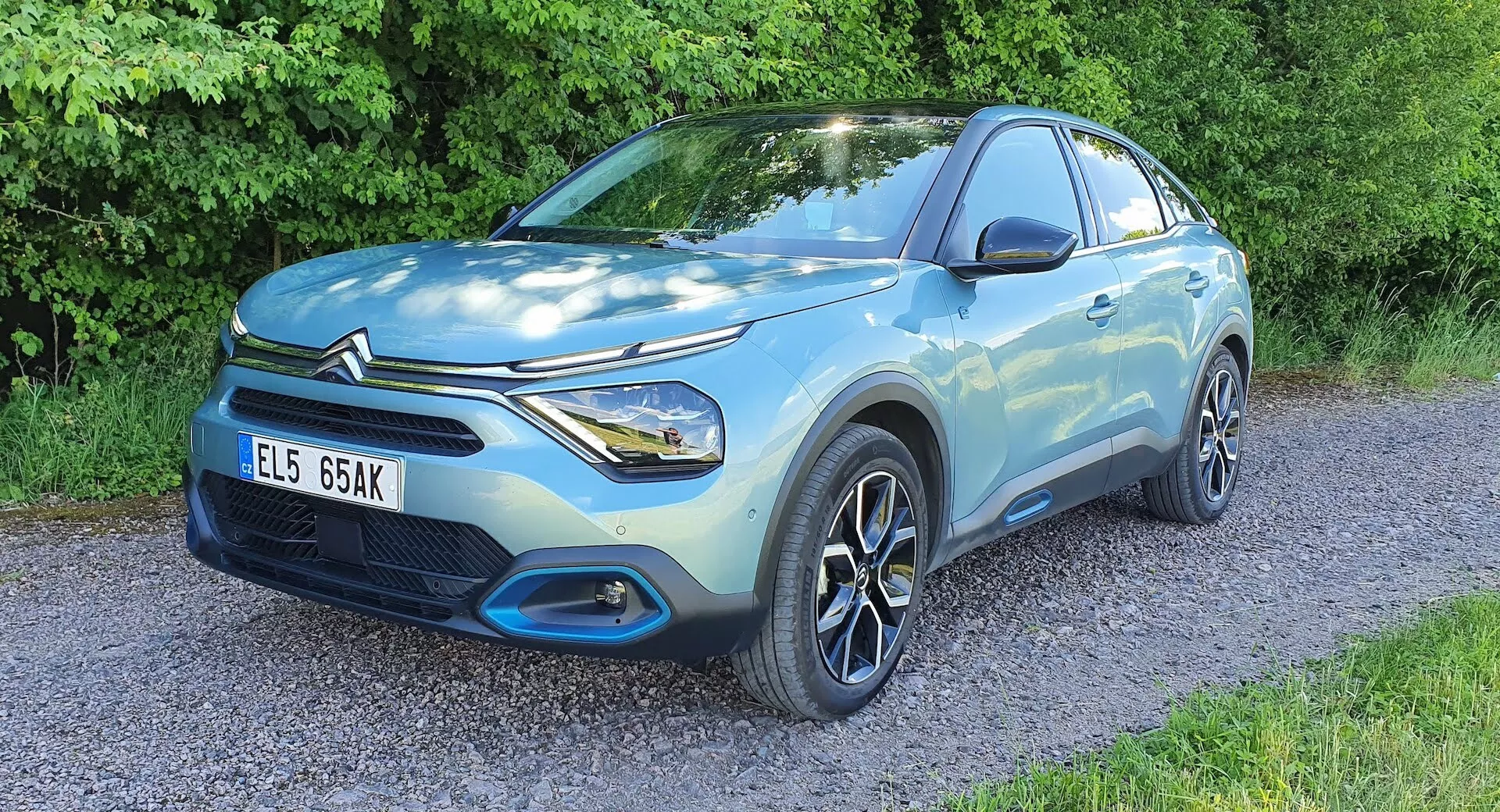 Világoskék Citroen C4 2020 (e-C4) kompakt SUV kavicsos felületen parkol, háttérben növényzettel, elülső negyed nézetben, amely a vezető oldalát, elülső homlokzatát, szoborszerű karosszériát, LED fényszórókat, többrudazatos alufelniket, első rácsot és a gyártó emblémáját mutatja, kiemelve a dizájn és technológiai jellemzőket.