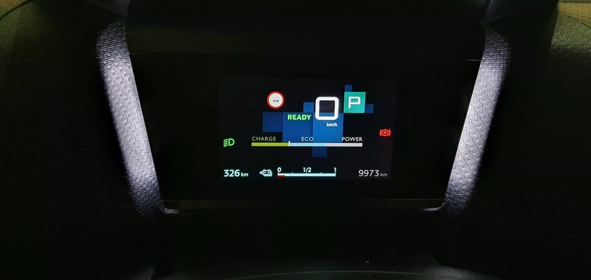 A Citroen C4 2020 (e-C4) digitális műszerfalának közeli nézete, amely 'CHARGE' és 'ECO' jelzőket mutat, 'READY' jelet, '32.6'-os akkumulátor ikont, környezetbarát levélikont, 'POWER' felhasználásjelzőt, 'P' parkolási szimbólumot, lefelé mutató nyíllal rendelkező '10' hőmérséklet kijelzőt, és '9973 km' kilométerszámlálót, amelyet a vezetőülés szemszögéből rögzítettek.