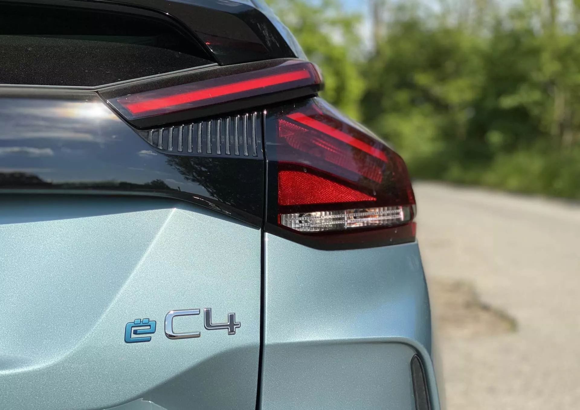 Közelről a Citroen C4 2020 (e-C4) bal hátsó oldala, részletes LED hátsó lámpával, csomagtartó szélével és a hátsó negyed panel egy részével; 'ë-C4' emblémával, amely az elektromos változatra utal; világos metallic szín fényes felülettel; sekély élesség mélységgel, elmosódott kültéri háttér zöld növényzettel.