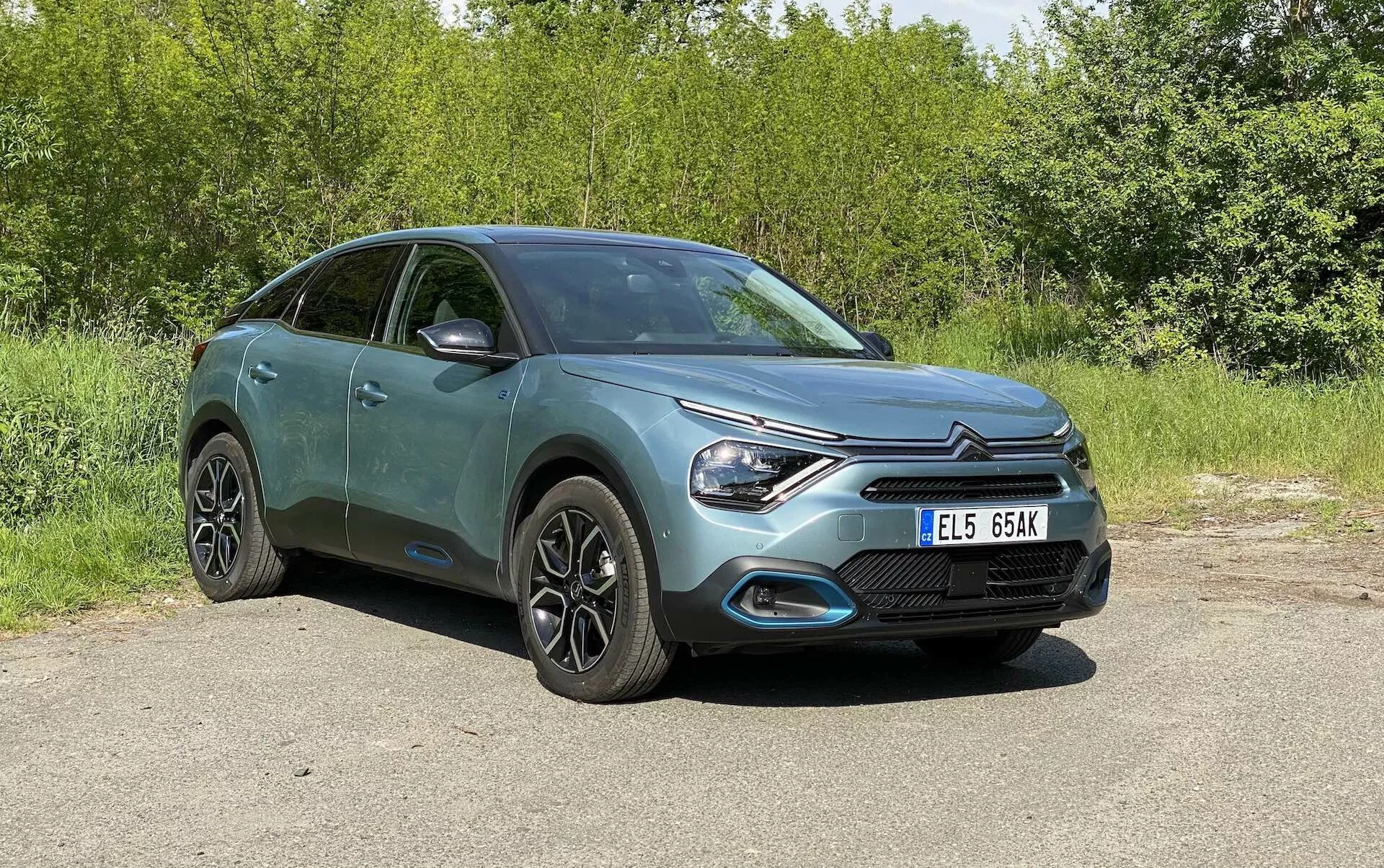 Egy Citroën C4 2020 (e-C4) teljes nézete parkolva, kívülről, elülső háromnegyed szögből fényképezve, megmutatva az első rácsot, az elegáns LED fényszóró dizájnt és a motorháztető formáját, valamint részben az oldalsó profilját, beleértve az ajtókat és oldalsó tükröket. A crossover/SUV kialakítású ferdehátú modern könnyűfém kerekekkel rendelkezik, integrált irányjelzőkkel a tükrökben és egy metálfényezéssel. A háttér zöldellő, és az autó természetes nappali fényben látható zavaró elemek nélkül.