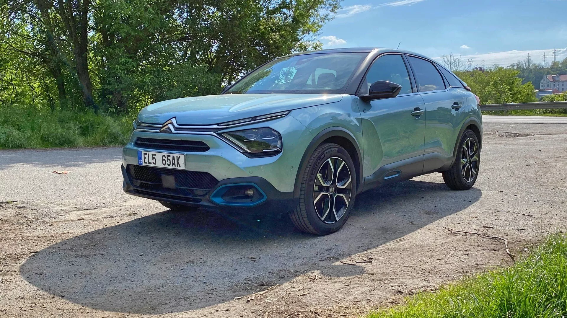 A képen egy 2020-as Citroen C4 (e-C4) hatchback vagy kompakt SUV alacsony előrébb-felé háromnegyedes szögből látható. Ez a perspektíva kiemeli az elülső részt, beleértve a hűtőrácsot, a fényszórókat és a lökhárítót, valamint a vezetőoldali profilt, amely magában foglalja az első sárvédőt, a kereket, a vezető ajtaját és a hátsó ajtó egy részét. Az autó modern, sportos dizájnnal rendelkezik, sima vonalakkal, lejtős tetővonallal, jellegzetes LED-es első fényszórókkal és egy kiemelkedő első ráccsal. Az elülső lökhárítón egy kontrasztos fekete díszítés található, amely növeli a robusztus megjelenését. A bonyolult többszörös küllős kerékdizájn a stílusra vagy a teljesítményre helyezi a hangsúlyt. A fényes metálfényezés tiszta és polírozott külsőt tükröz a napfényben. Az autó zöld környezetben parkol, amint azt a fák és a világos időjárási viszonyok is mutatják. A technológiai jellemzők, mint például az érzékelők vagy a kamerák, ebből a szögből nem igazolhatók, és a rendszámtábla látható, de az adatvédelmi okokból kihagyásra került.