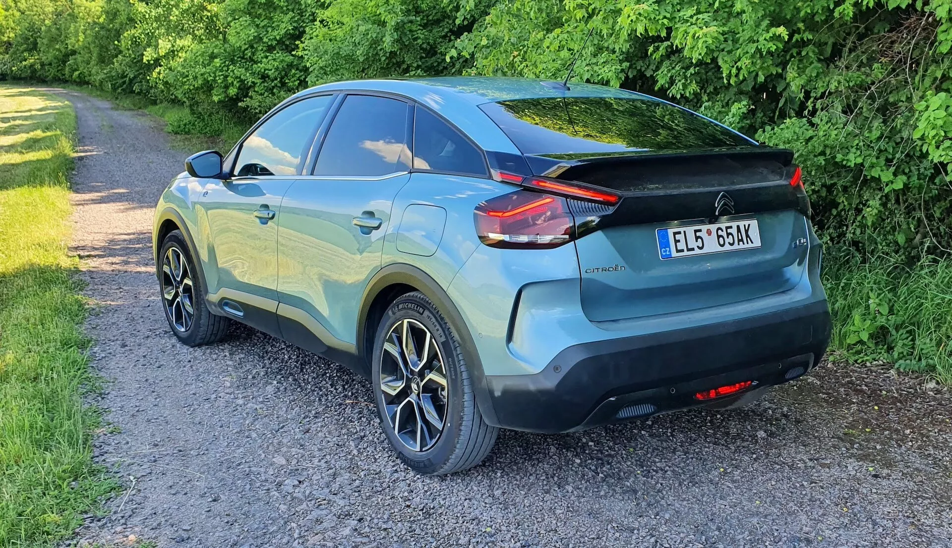 A képen egy Citroen C4 2020 (e-C4) látható háromnegyedes hátsó nézetből, amely bemutatja a modern crossover dizájnját, egy keskeny utakra parkolva, oldalt növényzettel. Az autó jellegzetes LED hátsó lámpákkal rendelkezik, amelyek a hátsó negyedpanelek felé nyúlnak, tető spoilerral, két tónusú festéssel és ötágú könnyűfém kerekekkel. Látható a hátsó ablaktörlő, az integrált hátsó ajtókilincsek és a hátsó lökhárítón lévő visszaverő elemek. Az autó kifogástalan állapotúnak tűnik, a világítás késő délutánt vagy estét sugall.