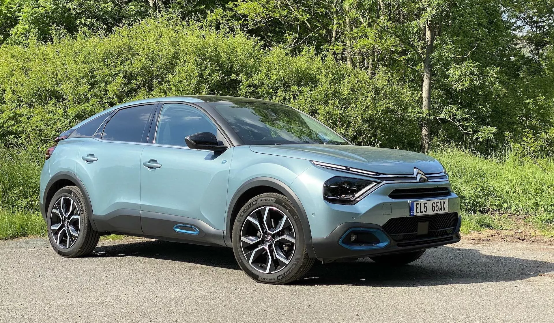 Alacsony szögű elölnézet egy 2020-as Citroen C4-ről (e-C4), amely szabadban parkol, természetes környezetben, növényzettel a háttérben. Modern, aerodinamikus kialakítás, jellegzetes első hűtőrács központi emblémával, keskeny LED fényszórók, fekete betétekbe integrált ködlámpák, többrészes könnyűfém keréktárcsák, fényes menta vagy világos türkizkék karosszériaszín, sima, kontúros vonalak a motorháztetőn és oldalpaneleken, integrált irányjelzőkkel ellátott visszapillantó tükrök, enyhén lejtős tetővonal, amely egy crossover SUV karosszériatípust sejtet, fekete burkolat a kerékívek körül és az autó alsó oldalán, valamint a szélvédő egy része, amely tükrözi a napsütéses időben az égbolt fényét.