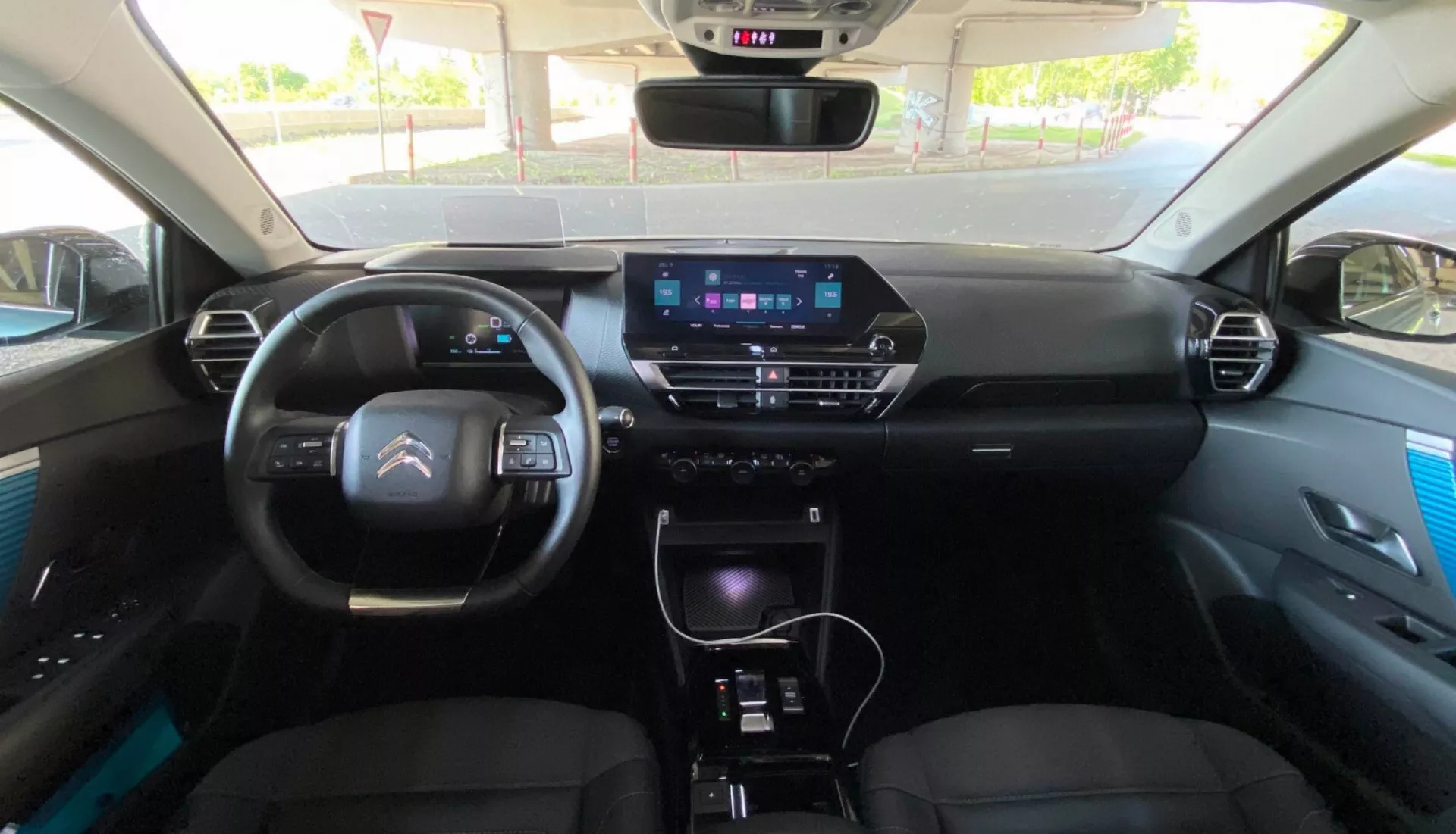 A Citroen C4 2020 (e-C4) belső nézete a hátsó ülésből, amely a műszerfalra fókuszál digitális műszerfallal, modern kormánykerékkel beépített vezérlőkkel és kimagasló információs képernyővel a középkonzolon. A konzol tartalmazza a klímavezérlő gombokat, a sebességváltókart és további vezérlőket, jelezve olyan funkciókat, mint a GPS navigáció, multimédia rendszer és kézi vagy automata klímavezérlés. Az első ülések kárpitozottak fejtámlákkal, és az ajtópanelek prémium elemeket mutatnak.