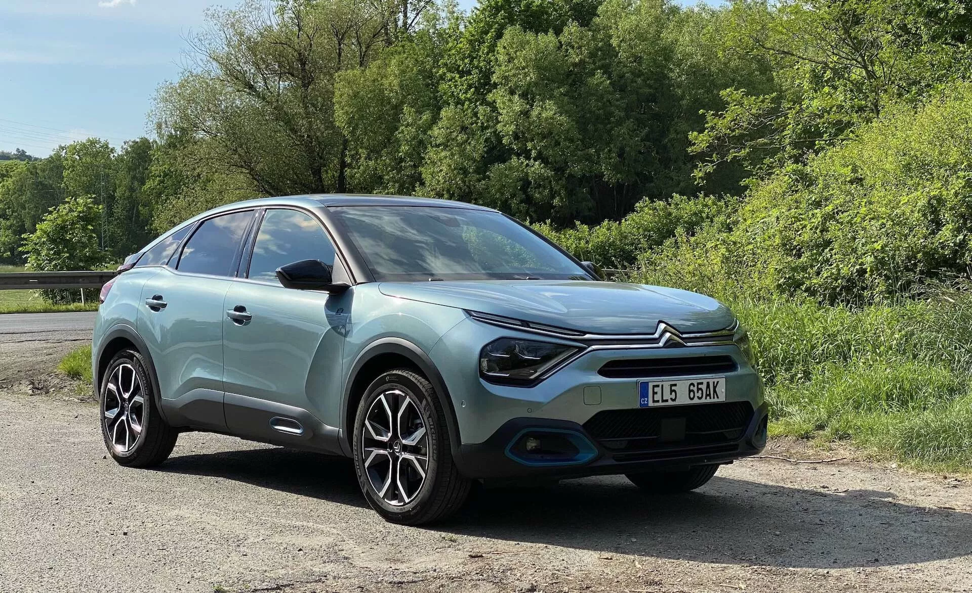 Egy Citroen C4 2020 (e-C4) képe háromnegyedes frontális perspektívából, kiemelve a pasztellkék karosszériát kontrasztos fekete tetővel, modern LED-fényszórókkal, formás első lökhárítóval, ötvözet keréktárcsákkal és összetett küllőmintázattal. A kép enyhén lefelé irányuló szögből készült, részletesen bemutatva a bal oldali fényszórót, az első lökhárítót, az első kereket, a szélvédőt és a vezetőoldali ablakot, a rendszámtáblával jól láthatóan elöl. Az autó buja zöld háttérben, vidéki vagy parkos környezetben, napfényes nappali világításban van elhelyezve.