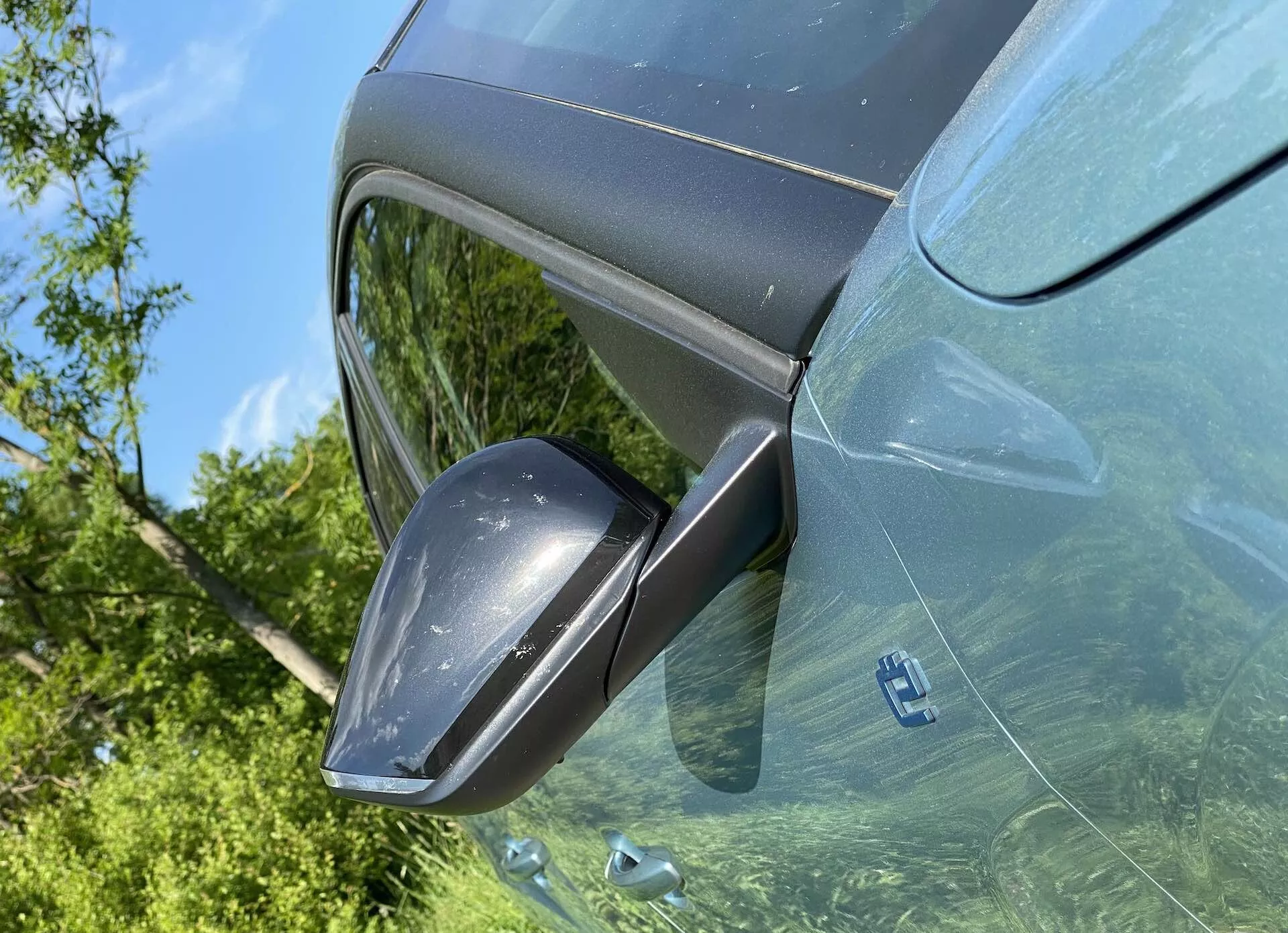 A Citroen C4 2020 (e-C4) vezetőoldalának közelije egy felfelé irányuló szögből, fókuszban a modern oldalsó visszapillantó tükörrel, integrált irányjelzőkkel, színben a karosszéria színéhez illeszkedő borítással, amely tükrözi a kültéri növényzetet a sötétített ablakokon. Világos nappali jelenet kiemeli a fémes festés és a jármű elegáns, modern formatervezését.