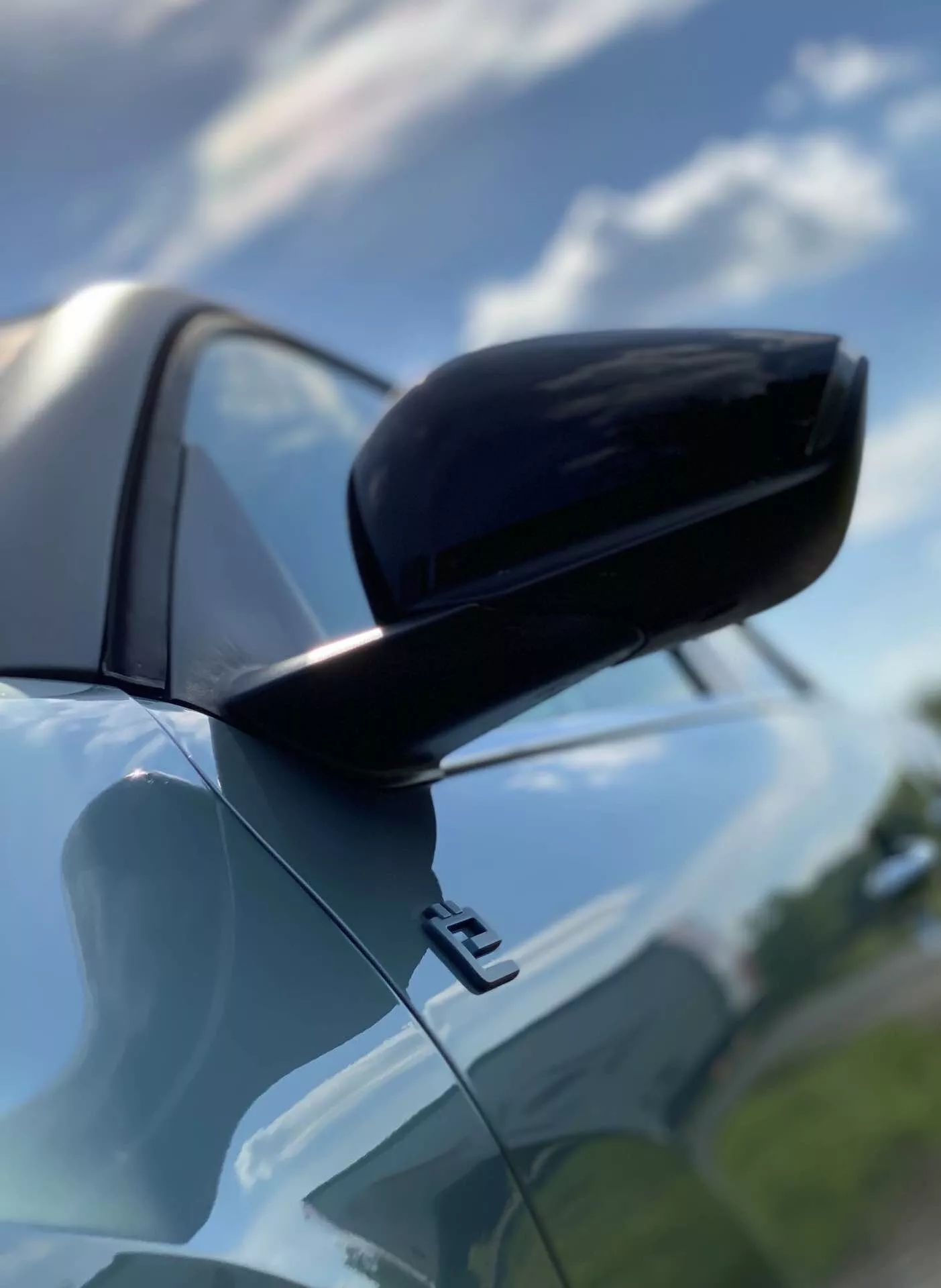 Közelkép a Citroen C4 2020 (e-C4) jobb oldali visszapillantó tükréről az utasoldalon, alacsony szögből nézve, amely bemutatja a karosszéria színével megegyező héjat fekete alsó résszel, kétszínű kialakításban. Kiemelkedik az aerodinamikus burkolat, valamint a fényes felület és a részben felhős égbolt tükröződését természetes nappali fényben. A lökhárító első része és az ajtó széle felfedi az autó kortárs dizájnját.