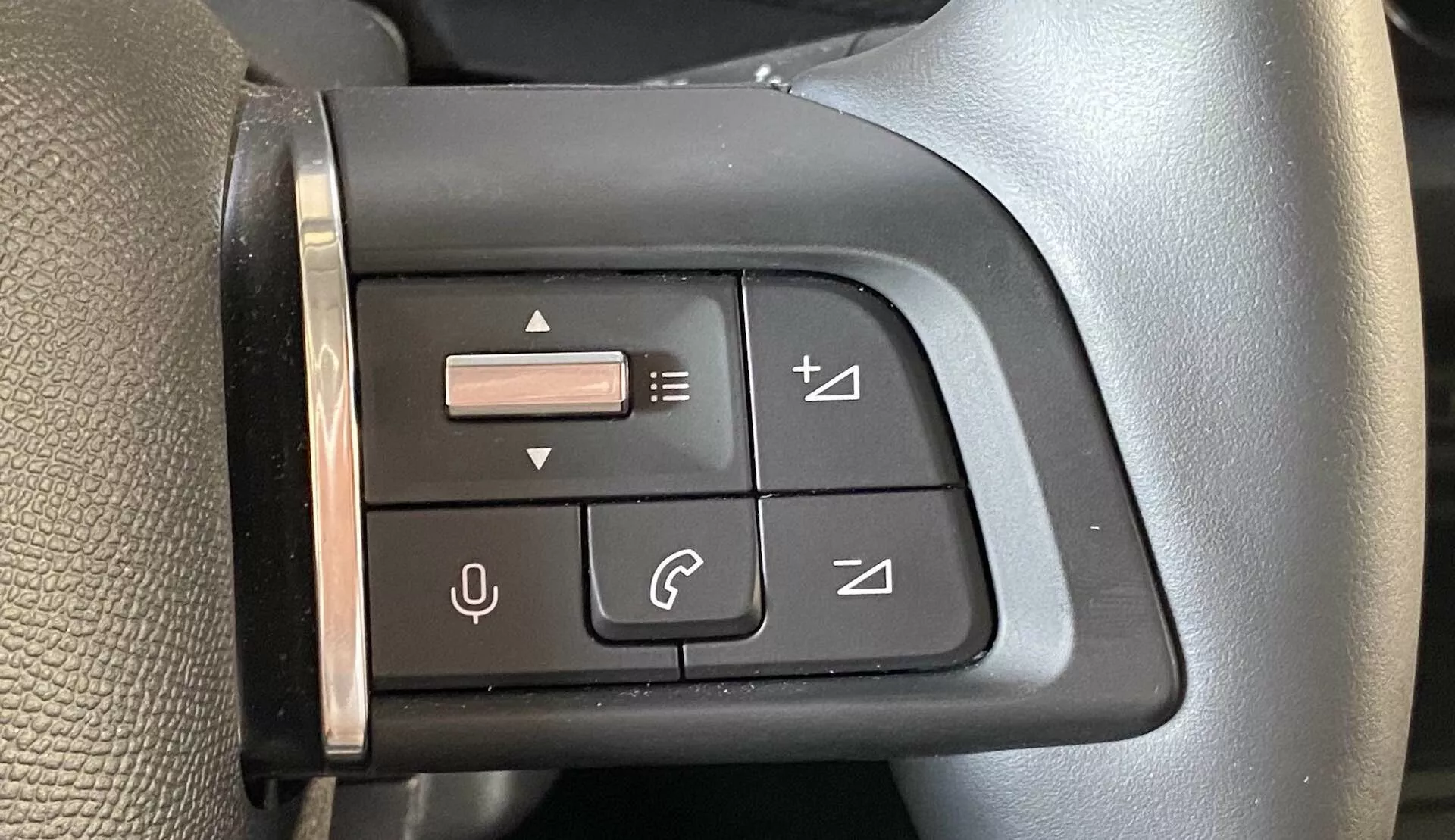 Közelkép a Citroën C4 2020 (e-C4) bal oldali kormánykerék multifunkciós vezérlőpaneljéről a vezető nézőpontjából. A panel gombokkal van ellátva hangerőszabályozáshoz, számváltáshoz, telefonhívások kezeléséhez, valamint egy menünavigációs joystickhoz. A fekete panel és a gombok tapintásra érzékenyek és illeszkednek a kormánykerék esztétikájához, amely texturált felülettel és nagy minőségű anyagra utaló varrással rendelkezik.