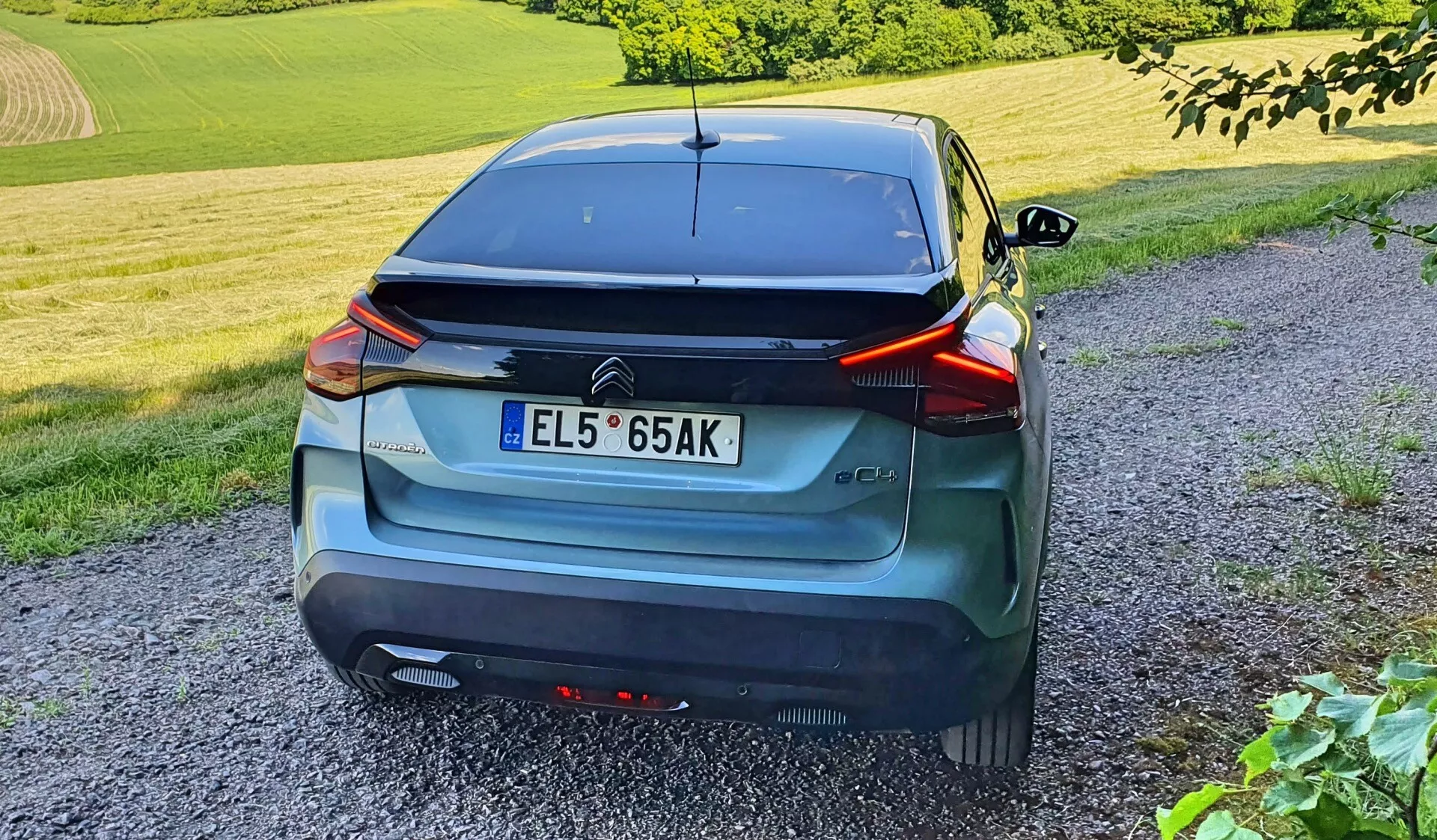 Egy modern hatchback autó hátulnézete háromnegyedes szögből megörökítve, amely kiemeli elegáns vonalait, körbefutó hátsó lámpáit, központi rendszámtábla helyezését, aerodinamikus spoilert és hátsó ablaktörlőt. A jármű szürke vagy kék metál színű, és lökhárítón található érzékelőkkel van felszerelve. Az autó egy kavicsos úton parkol falusias környezetben, fű és lomb mellett, késő délután vagy kora este fényviszonyai mérhetők fel, álló ember magasságából fényképezve.
