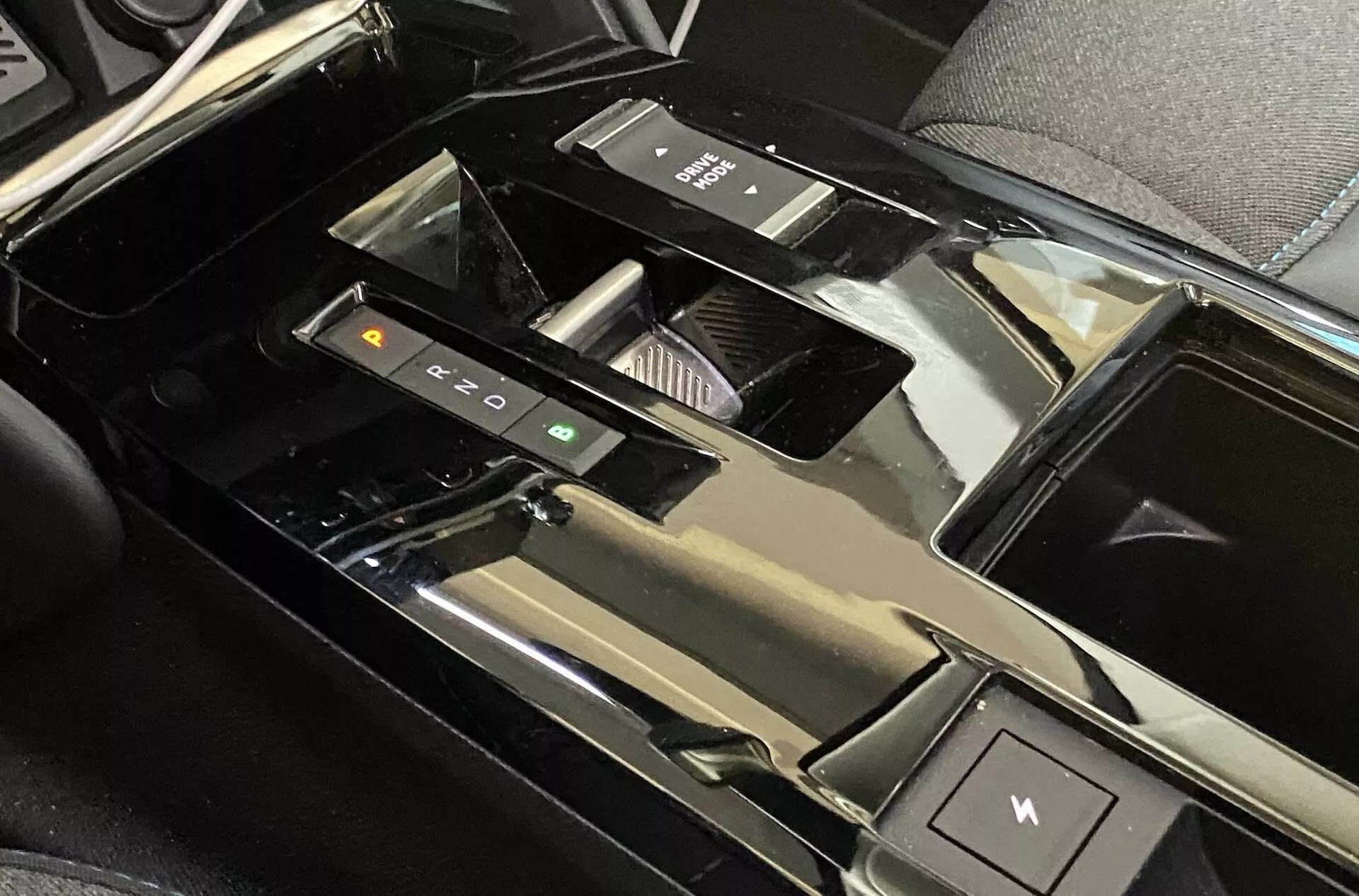 Közelkép a Citroën C4 2020 (e-C4) középkonzoljáról, kiemelve az elektronikus sebességváltót, a vezetési mód vezérlő gombokat, a pohártartókat, a fényes tárolórekesz fedelét, az elektronikus kézifékkapcsolót, a fényes fekete anyagbevonatot és a részben látható érintőkijelzős felületet, ami egy magas technológiai szintű, luxus járműbelsőre utal.