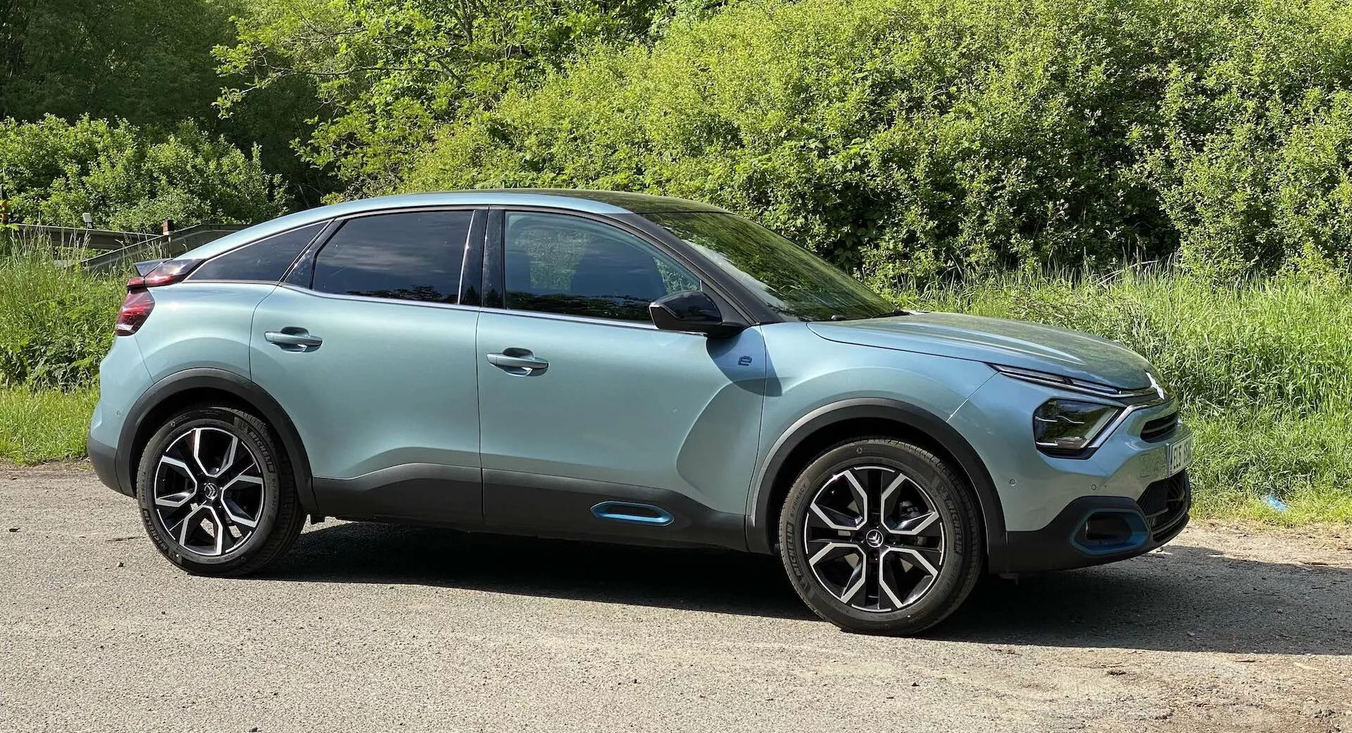 Citroen C4 2020 (e-C4) modern crossover SUV oldalsó profilképe alacsony szögből, enyhén földszint felett fényképezve. Az autó sík útszéli felületen parkol, háttérben zazöld növényekkel, ami vidéki vagy parkos környezetre utal. A karosszéria elegáns motorházteteje ívelt első rácshoz vezet, LED nappali menetfényekkel és potenciálisan ködlámpákkal. A kocsitest kétszínű, világos türkiz vagy menta zöld, fekete díszítéssel a kerékcsapok körül és az alsó oldalsó szoknya részen. Ez a színséma kiterjed az oldalsó tükrökre is, amelyekbe beépített irányjelzők találhatók – másik biztonsági és technológiai szempont. A karosszéria színére festett ajtókilincseken találhatók valószínűleg kulcsnélküli beléptető érzékelők. A nagy, sokküllős könnyűfém kerekek és a nagyteljesítményű gumik az esztétikát és a teljesítményt hangsúlyozzák. Az autó tetővonala enyhén lejt a hátsó rész felé, ami sportos vagy aerodinamikai profilt sugall. Az ablakfelületek sötétített üveg borítással rendelkeznek, amely UV-védelmet és diszkréciót nyújthat az utasoknak, így szintén technológiai előnyt jelent a komfort és biztonság szempontjából. A technológiai fronton ez a jármű feltehetőleg fejlett vezetői segédrendszereket (ADAS) tartalmaz, beleértve a sávelhagyás figyelmeztetést, az adaptív sebességtartó automatát és a parkolási asszisztenst, amely szenzorok jelenlétével bizonyítható – bár ezek nem kifejezetten láthatóak a képen. Az képet napfénynél, jó láthatóság mellett készítették, és az autó közepes távolságra van a kamerától, lehetővé téve a jármű oldalsó profiljának teljes nézetét. A szög és a keretezés a jármű dizájnjának és külső jellemzőinek standard bemutatásának kategóriájába sorolható.