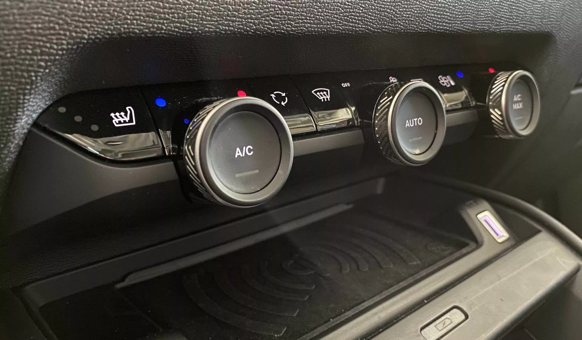 Közelképen a 2020-as Citroën C4 (e-C4) klímavezérlő rendszere látható, három kör alakú kezelőgombbal a középkonzolon, enyhén felfelé döntött szögből. A bal oldali gomb a fűtött üléseket szabályozza kék hőmérsékleti szintjelzőkkel, a középső gomb 'A/C' felirattal légkondicionáláshoz, a jobb oldali gomb pedig 'AUTO' felirattal az automatikus klímabeállításokhoz. A kék és piros jelzők a hőmérsékleti beállításokat jelzik. A gombok alatt egy rekesz található vezeték nélküli töltőlappal, amely rádióhullámokat szimbolizáló jelet tartalmaz, amelyet matt fekete műanyag burkolat és mintás textúra vesz körül.