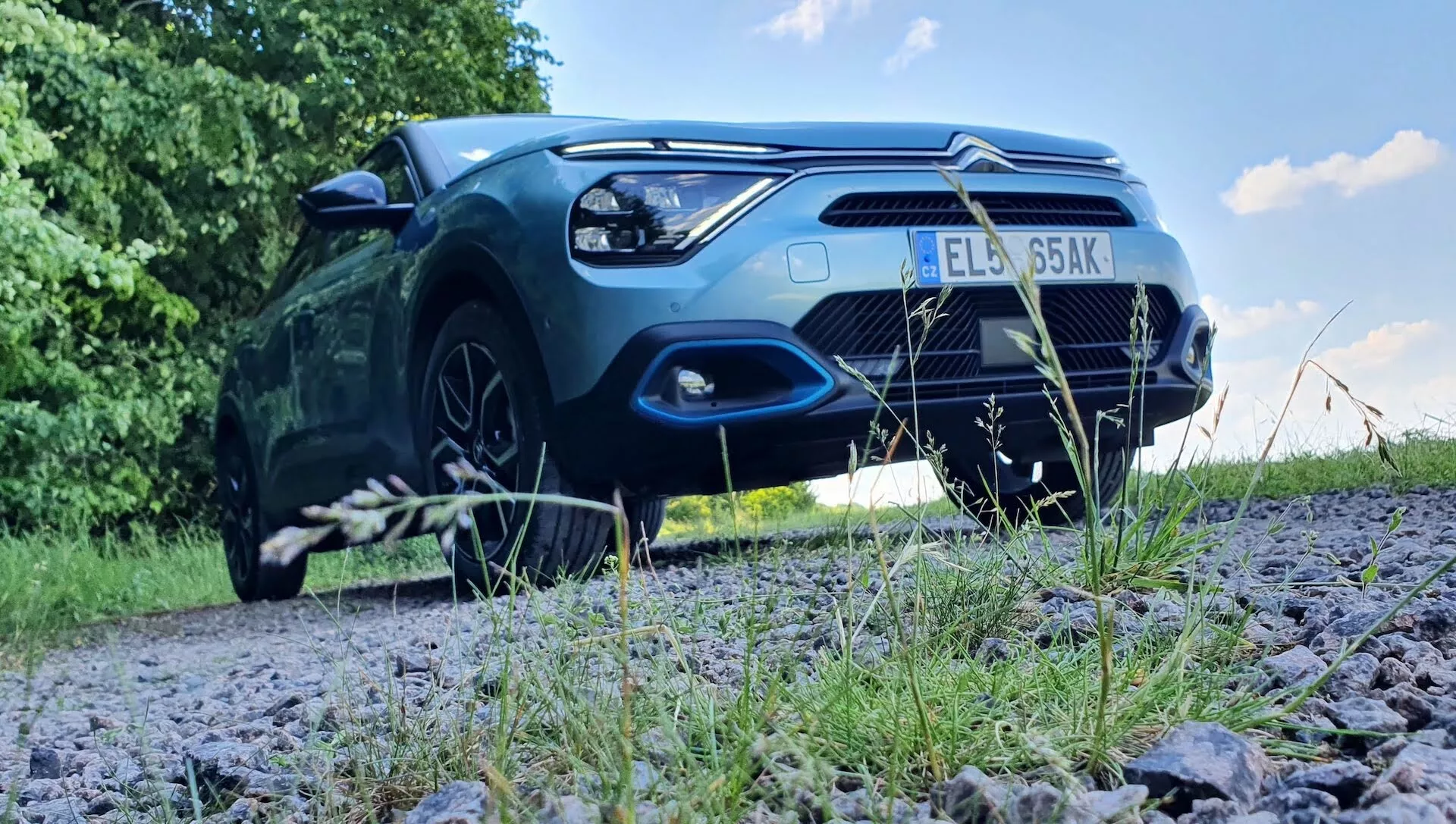 Alacsony szögű frontális nézet egy parkoló Citroën C4 2020-as (e-C4) autóról, amely egy kavicsos úton áll, zöld környezetben. Látható a motorháztető, hűtőrács, első lökhárító, fényszórók és az első kerekek egy része. A kép kiemeli a jármű robusztus kiállását és külső design jellemzőit, mint például a fényszórók formáját, a hűtőrács mintázatát, a légbeömlő nyílásokat és a motorháztető kontúrjait. A fényezésen tükröződő napfény fokozza az autó esztétikai vonzerejét. A lehetséges fejlett világítási technológiák és parkolószenzorok is a látható technológiai jellemzők közé tartozhatnak.