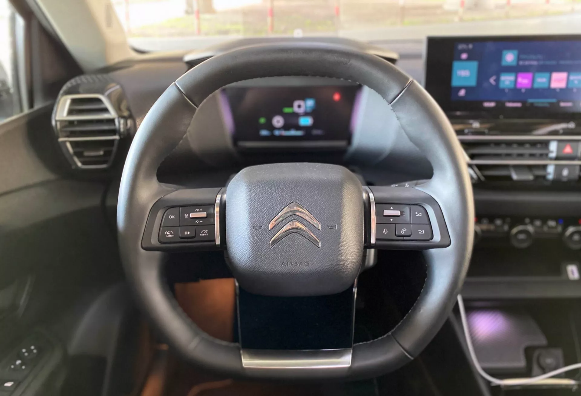 A vezető vagy az első utas szemszögéből készített belső nézet a 2020-as Citroën C4 (e-C4) modellről, amely a kormánykereket és a modern műszerfalat mutatja. A háromküllős, bőrborítású kormánykerék hangvezérlésre, hangparancsokra és sebességtartó automatikára szerelt gombokkal rendelkezik. A digitális műszerfal színes kijelzőkkel és információs megjelenítéssel rendelkezik, amely fejlett digitális technológiára utal. Láthatók az infotainment érintőképernyőjének részei, a klímavezérlő gombok és a műszerfal fa hatású díszítése, ami kiemeli az autó technológiailag fejlett és esztétikailag kellemes belső kialakítását.