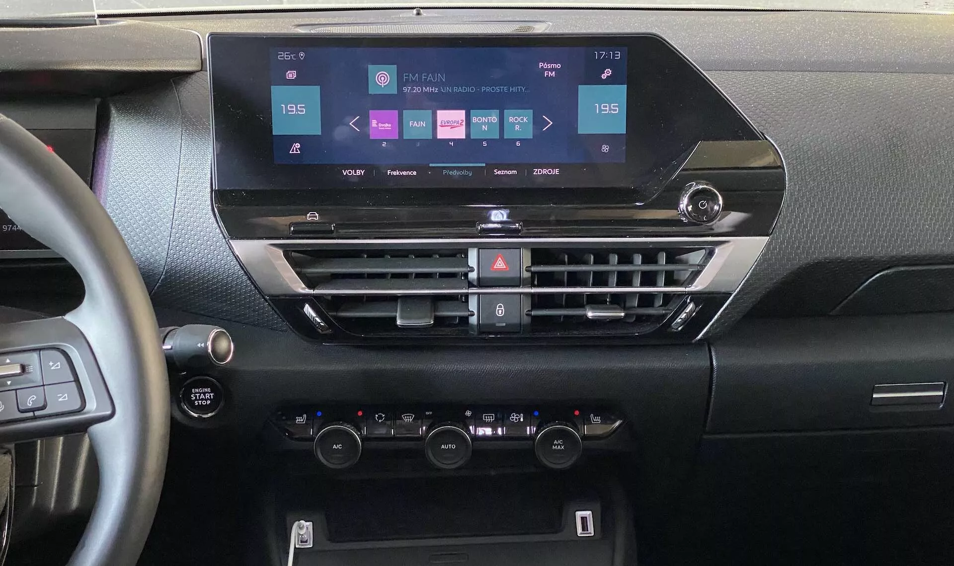 Citroen e-C4 2020 belső nézet, amely a műszerfalra összpontosít egy központi pozícióból. A képen egy modern érintőképernyős infotainment kijelző látható, navigáció, média, telefonkapcsolat és autóbeállítások opcióival. Az alatt vízszintes légbeömlők találhatók gombokkal a légkondicionálás és egyéb funkciók vezérlésére, majd duál zónás klímavezérlők hőmérséklet szabályzókkal és LCD kijelzővel. Látható a kormánykerék egy része beépített vezérlőkkel és egy start-stop gombbal, kiemelve a modern autótechnológiát és az ergonómikus kialakítást.