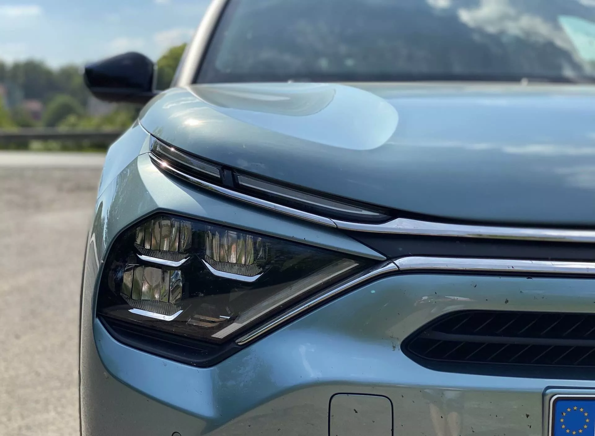 Közelképből, alacsony szögből készített felvétel egy modern jármű, konkrétan a Citroën C4 2020 (e-C4) bal első negyedéről. A fókuszban az elegáns, szögletes kialakítású fényszóró LED elemei láthatók, továbbá a horizontális lamellájú hűtőrács egy része, amely valószínűleg aktív zsalurendszerrel rendelkezik, valamint a lökhárítón elhelyezett parkolószenzorok láthatók. A kép a visszapillantó tükör egyes részeit is rögzíti, mely irányjelzőkkel vagy vakfoltfigyelő érzékelőkkel van felszerelve. A motorháztető sima kontúrja felemelkedő középső résszel rendelkezik, amely finom sportos karaktert kölcsönöz az autónak. A jól karbantartott, fényvisszaverő fényezés a kiváló minőségű anyagra vagy hatékony védőbevonatra utal. Az európai rendszámtábla részben látható, hangsúlyozva a modern autóipari technológiát és dizájnt.