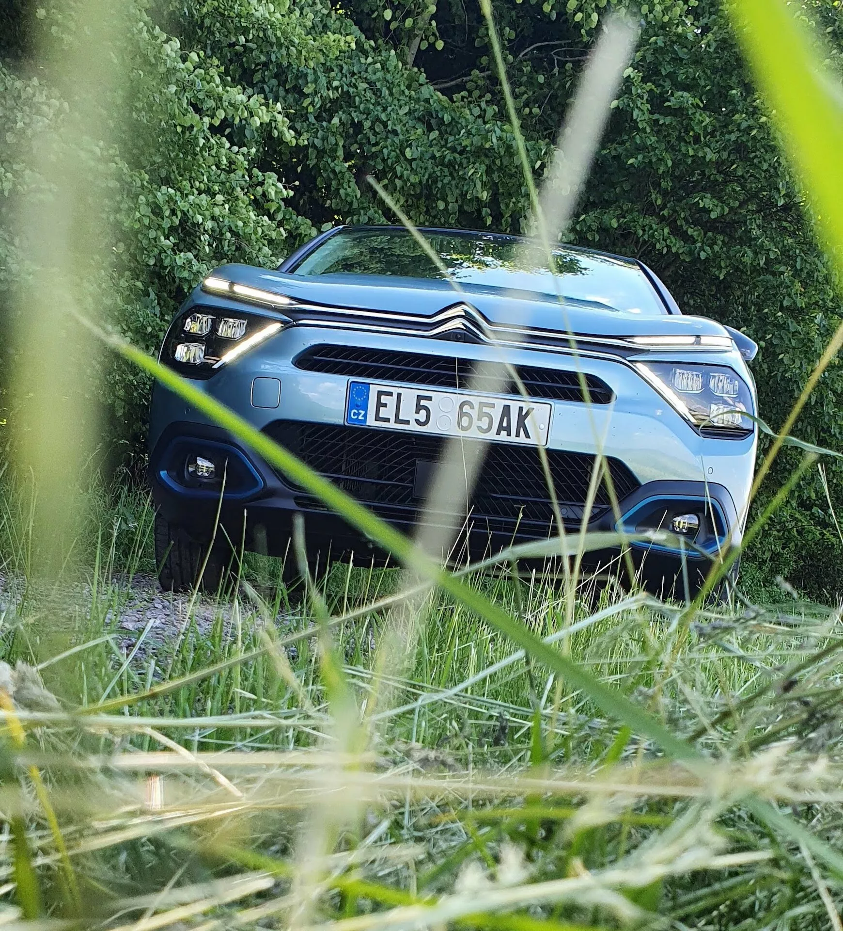 Egy kék Citroën C4 2020 (e-C4) autó, amelyen jól látható a dupla chevron embléma az első hűtőrácson, alacsony frontról nézve, zöld lombozattal keretezett háttér előtt. A kép kiemeli a jármű modern fényszóróit, elegáns első részét és a motorháztető középső, megemelt szakaszát. A fénykép kis mélységélessége élesen fókuszál az autóra, miközben kissé elhomályosítja a közvetlen környezetét, kiemelve a jármű dizájnját és technológiai jellemzőit.