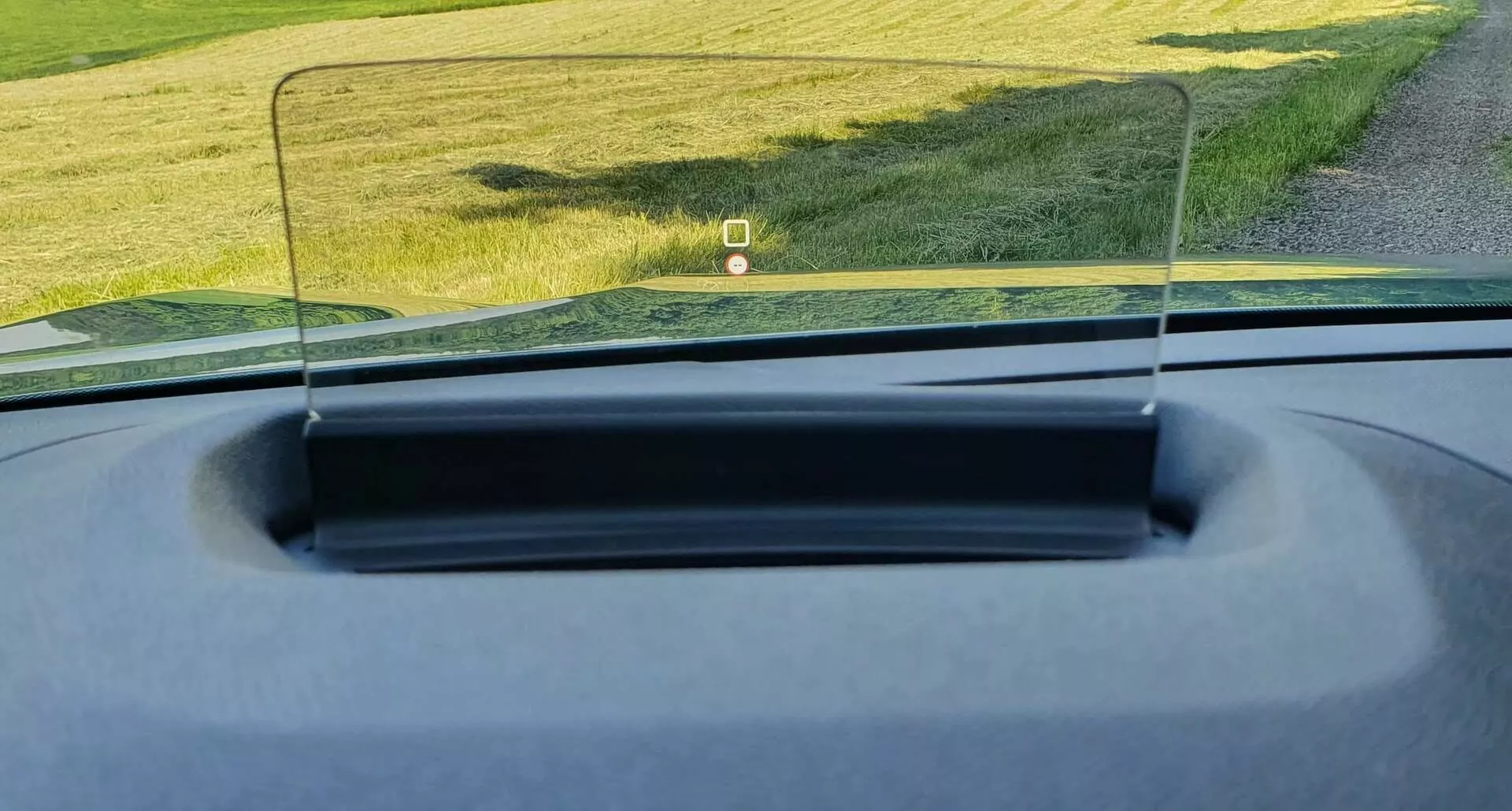 A Citroën C4 2020 (e-C4) belső terének perspektívája, amely a műszerfalat és a motorháztetőt mutatja egy head-up display-jel, amely információkat vetít egy átlátszó, téglalap alakú üvegfelületre. A fénykép a vezető vagy az utas üléséből készült, kiemelve a HUD-ot, mint fejlett vezetői asszisztens rendszert. Az autó látszólag egy vidéki területen parkol, ahol a háttérben zöld fű látható.