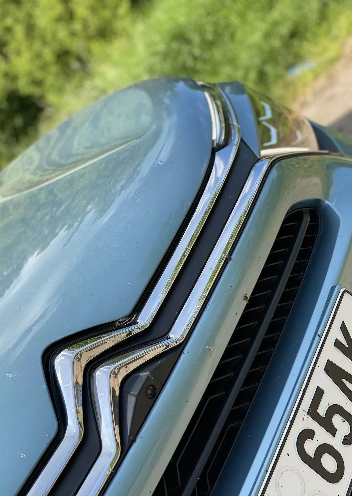 Autó közeli képe elölről és kissé felülről, kiemelve a függőlegesen orientált króm elemmel rendelkező első hűtőrácsot és vízszintes lamellákat, a felül elhelyezkedő márka emblémát, valamint az ívelt vonalakkal rendelkező motorháztető dizájnt. A motorháztetőn látható szennyeződés vagy pollen kültéri környezetre utal.