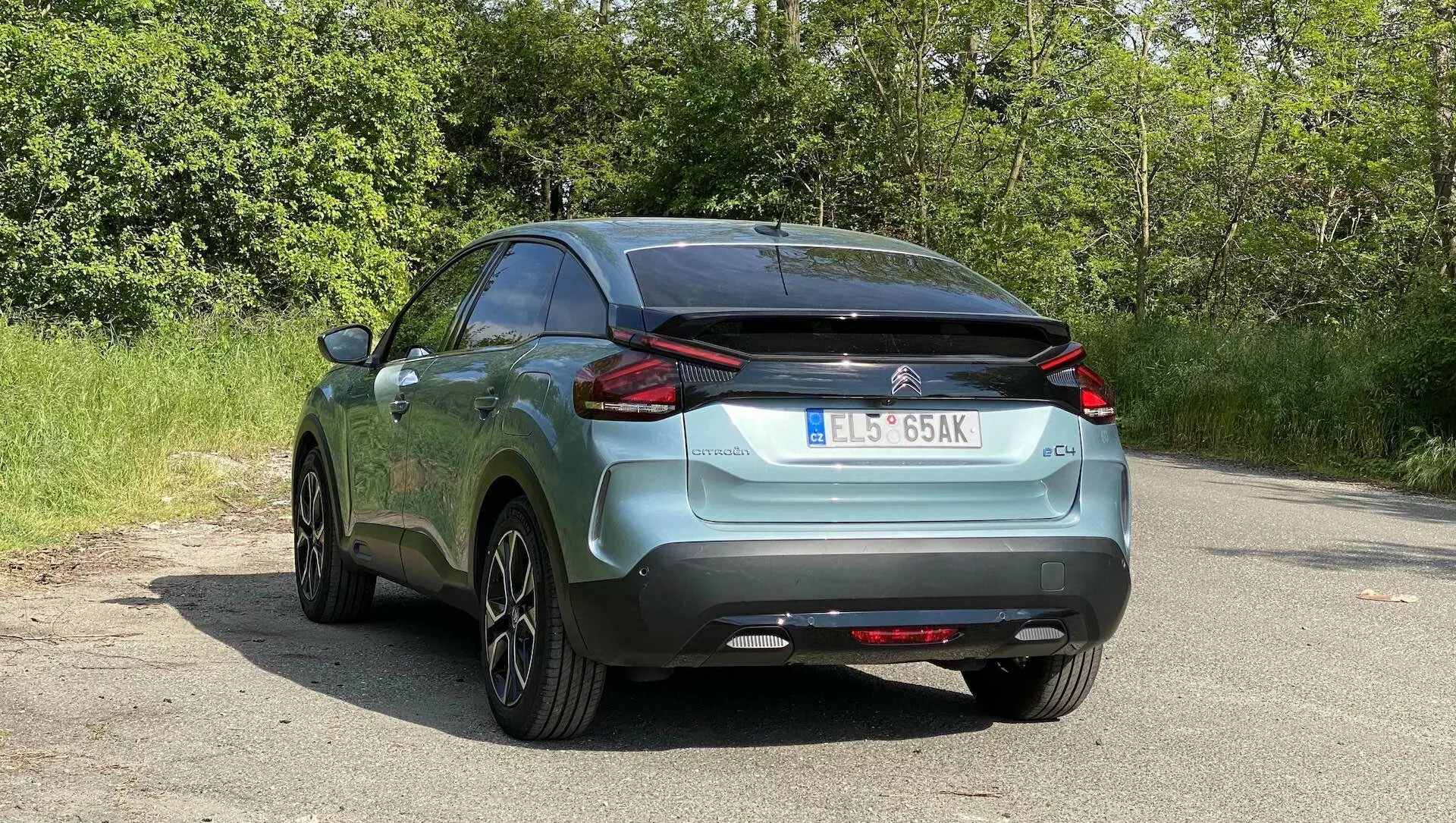 A 2020-as (e-C4) Citroen C4 hátsó szögből készült képe egy aszfaltos felületen parkolva, természetes lombozattal a háttérben, bemutatva a jellegzetes LED hátsó lámpákat, a márka- és modellszimbólumokat, a hátsó szélvédőt törlővel, a kéttónusú lökhárítót diffúzorszerű elemmel, beépített parkolószenzorokat, részben látható kipufogót, a tetőre szerelt antennát, többküllős alufelniket, a metálfényezést és az európai stílusú rendszámtáblát.