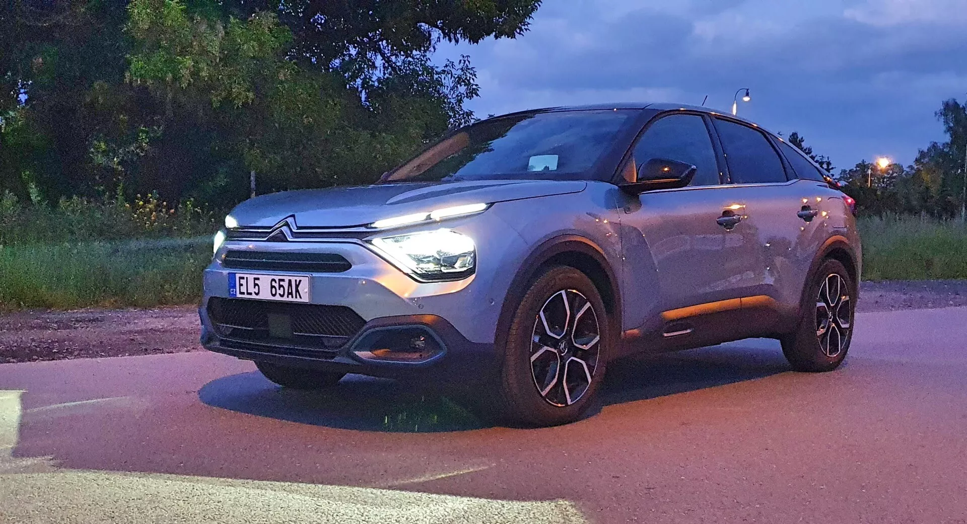 Egy Citroen C4 (e-C4) 2020-as modern crossover SUV szürkületkor, világító fényszórókkal és nappali menetfényekkel. Az autó háromnegyed elölnézetben látható, bemutatva az első homlokzatot, a márkajelzéssel ellátott hűtőrácsot, a motorháztetőt, az első lökhárítót és a vezető oldali profilt, beleértve a nagy, többküllős alufelniket. A jármű jelentős részét elfoglalja a képnek, bemutatva a LED fényszórókat, a jellegzetes hűtőrácsot, amely esetleg szenzorokat integrál, valamint a fémes festésű felületet, ami magasabb felszereltségi szinteket vagy testreszabási lehetőségeket sugall.