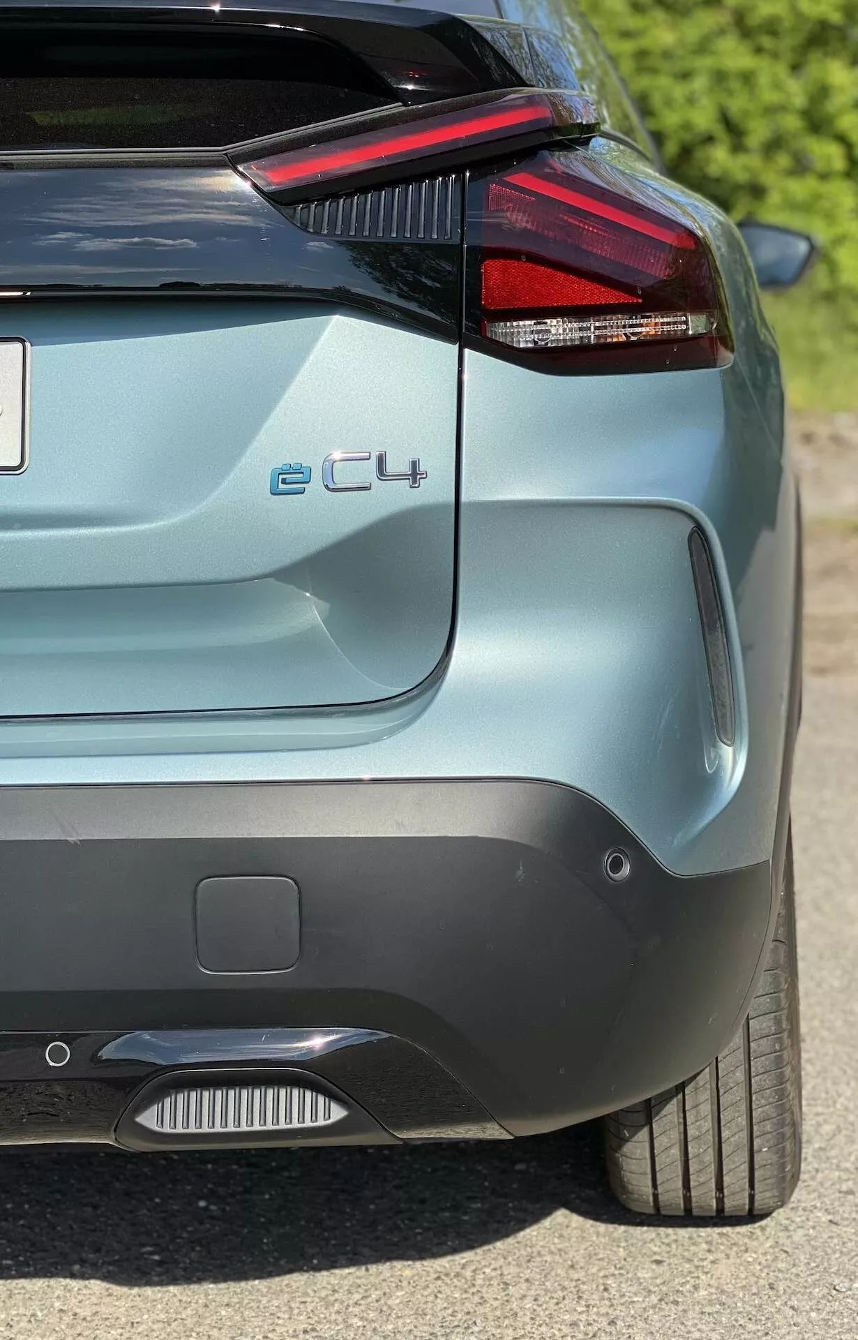 Hátsó nézet egy Citroën e-C4 2020-ról, amelyen látható a karakterisztikus, élesen megtervezett piros LED-es hátsó lámpatest, amely beleolvad a hátsó szélvédőbe, parkolásérzékelők a lökhárítón, a hátsó diffúzor egy része, a 'e-C4' modell jelvény, a stílusos alufelni és a metálfényezés. A kép a jármű jobb hátsó negyedére összpontosít felülről és oldalról.