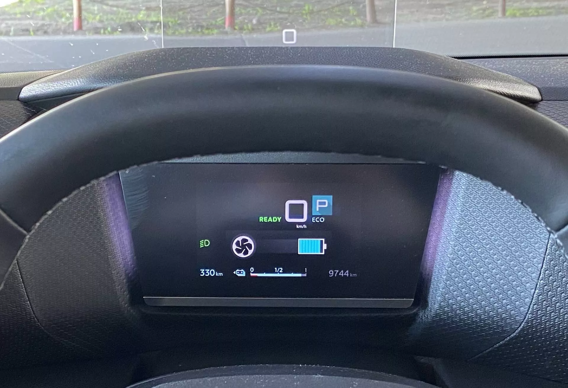 Közelről, a vezetőülésből készült nézet egy Citroën C4 2020 (e-C4) belsejéről, amely a fekete, texturált kormánykereket és a digitális műszercsoportot mutatja. A kormánykerék kissé kitakarja a digitális kijelző alját, amely számos funkciót jelez: 'READY' EV készenléti ikon, 'P' parkoló mód, 'ECO' mód, majdnem teljesen feltöltött akkumulátor ikon, '330' hatótávolság kijelző, 9744 km-es odaó, '+12°C' külső hőmérséklet és kerek nyíl ikon, amely valószínűleg a regeneratív fékezés erejének beállítására szolgál.