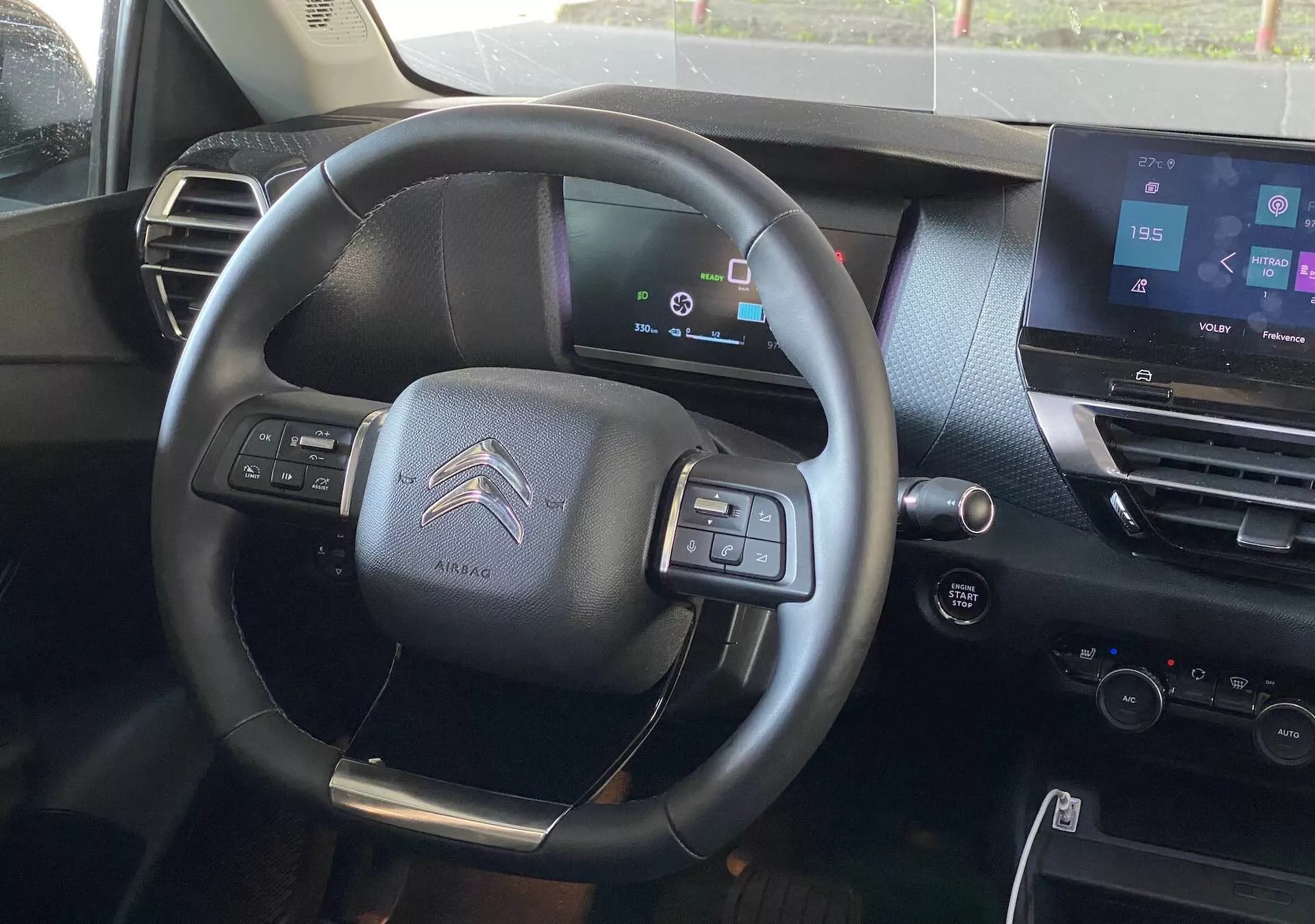 A Citroën C4 2020 (e-C4) belső nézete a vezetőülésből, középen egy háromküllős multifunkcionális kormánykerékkel, légzsák jelöléssel, digitális műszerfallal, amely a jármű állapotait mutatja, érintőképernyős infotainment rendszerrel a középkonzolon, sötét tónusú belső tervezéssel, audio és telefonos csatlakozási kezelőgombokkal, félautomata váltót sugalló váltófülekkel és a vezető ajtóján található elektromos ablakvezérléssel és tükörállítási lehetőségekkel.